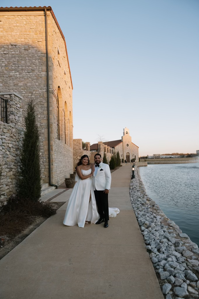 Couple dressed in wedding attire standing on a pathway by a body of water with stone buildings in the background during sunset.