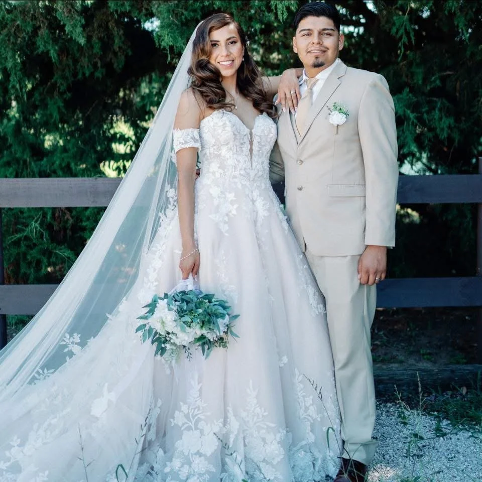 Bride and groom standing outdoors with green trees in the background, smiling for their wedding photo. The bride is wearing a white lace wedding gown with a long veil and holding a bouquet, while the groom is dressed in a cream suit with a boutonnièr