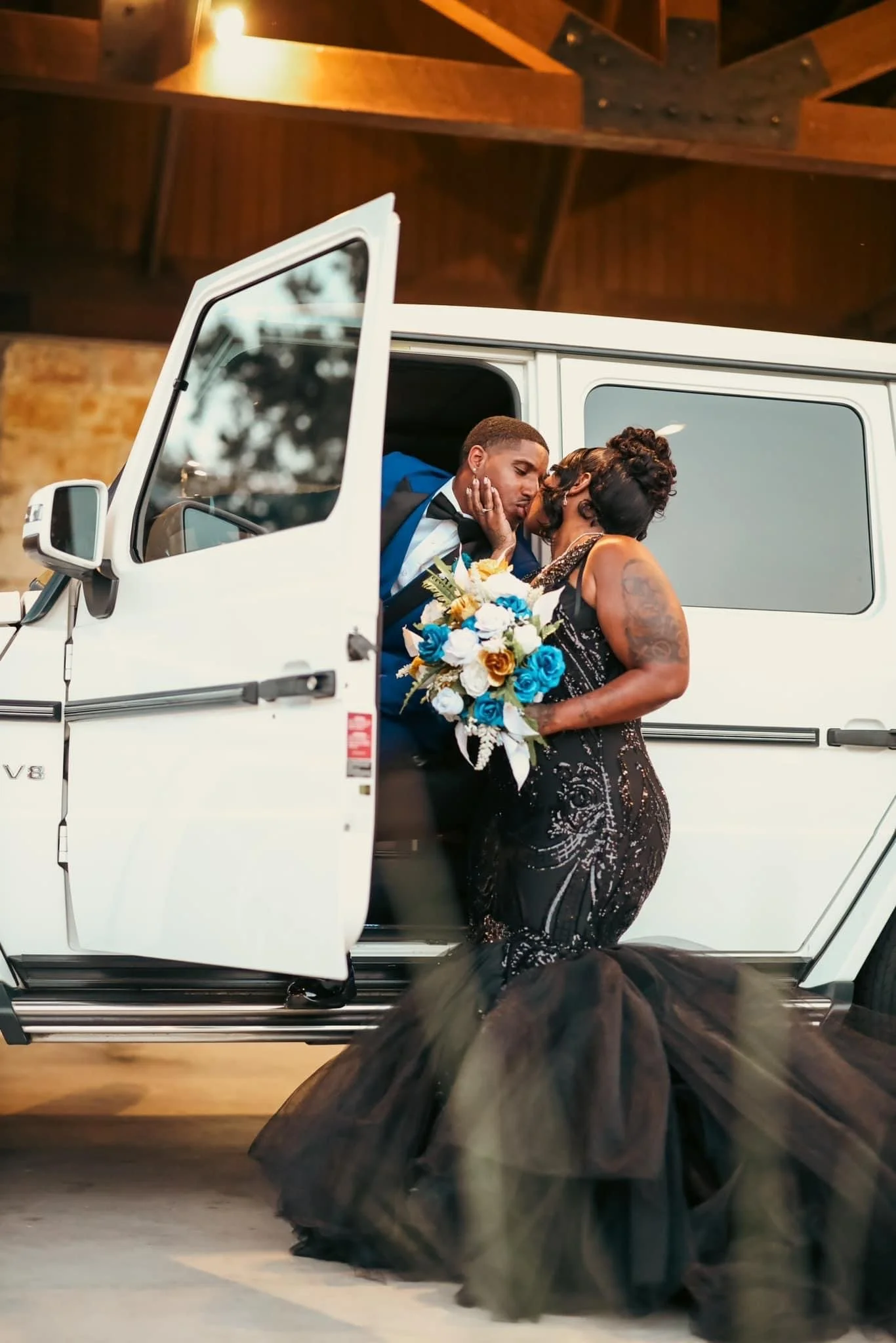 A couple dressed in formal attire sharing a kiss inside a white luxury car. The woman is holding a bouquet of blue, white, and gold flowers, and the man is wearing a tuxedo. The scene appears to be at a wedding or special event, with the car's door o