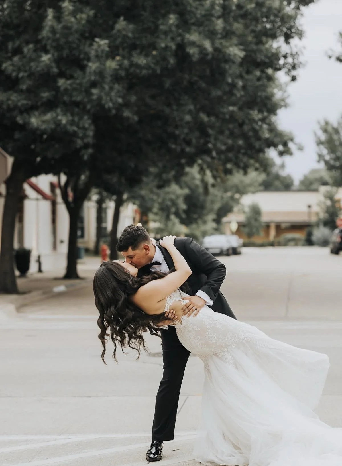 A newlywed couple sharing a kiss outdoors, with the groom dipping the bride in a street with trees and parked cars in the background.