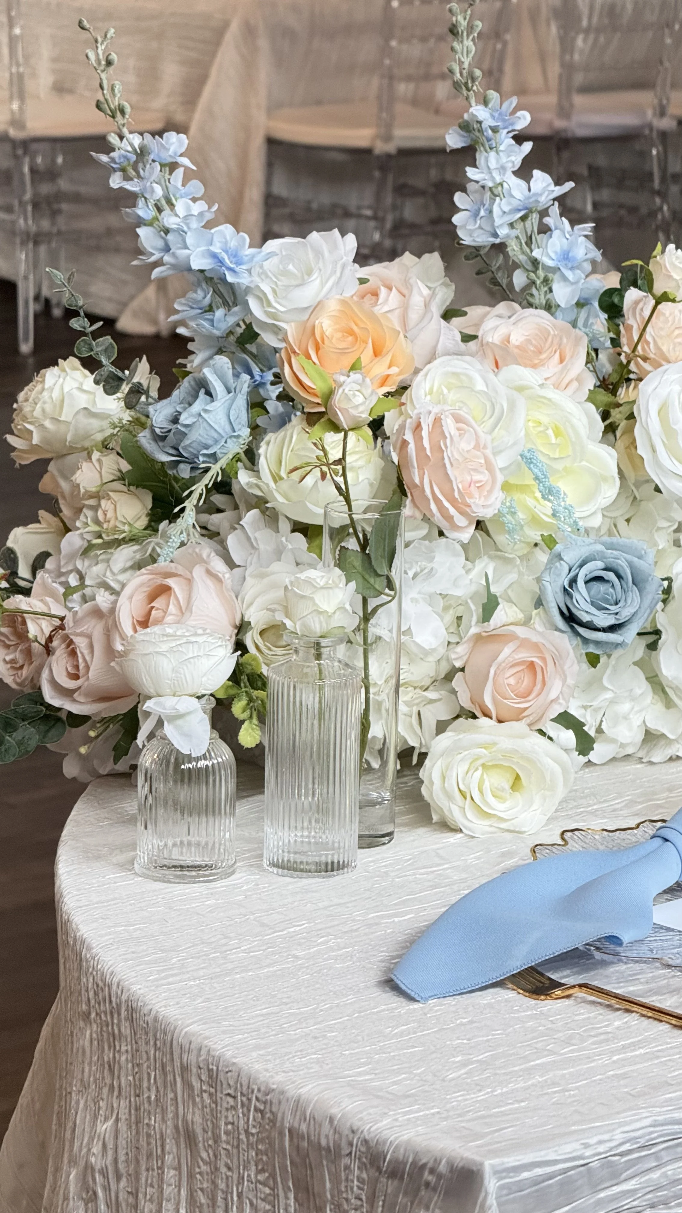 Elegant table with a large floral centerpiece of white, peach, and blue roses, accented with greenery, in clear glass vases, set for a special event.