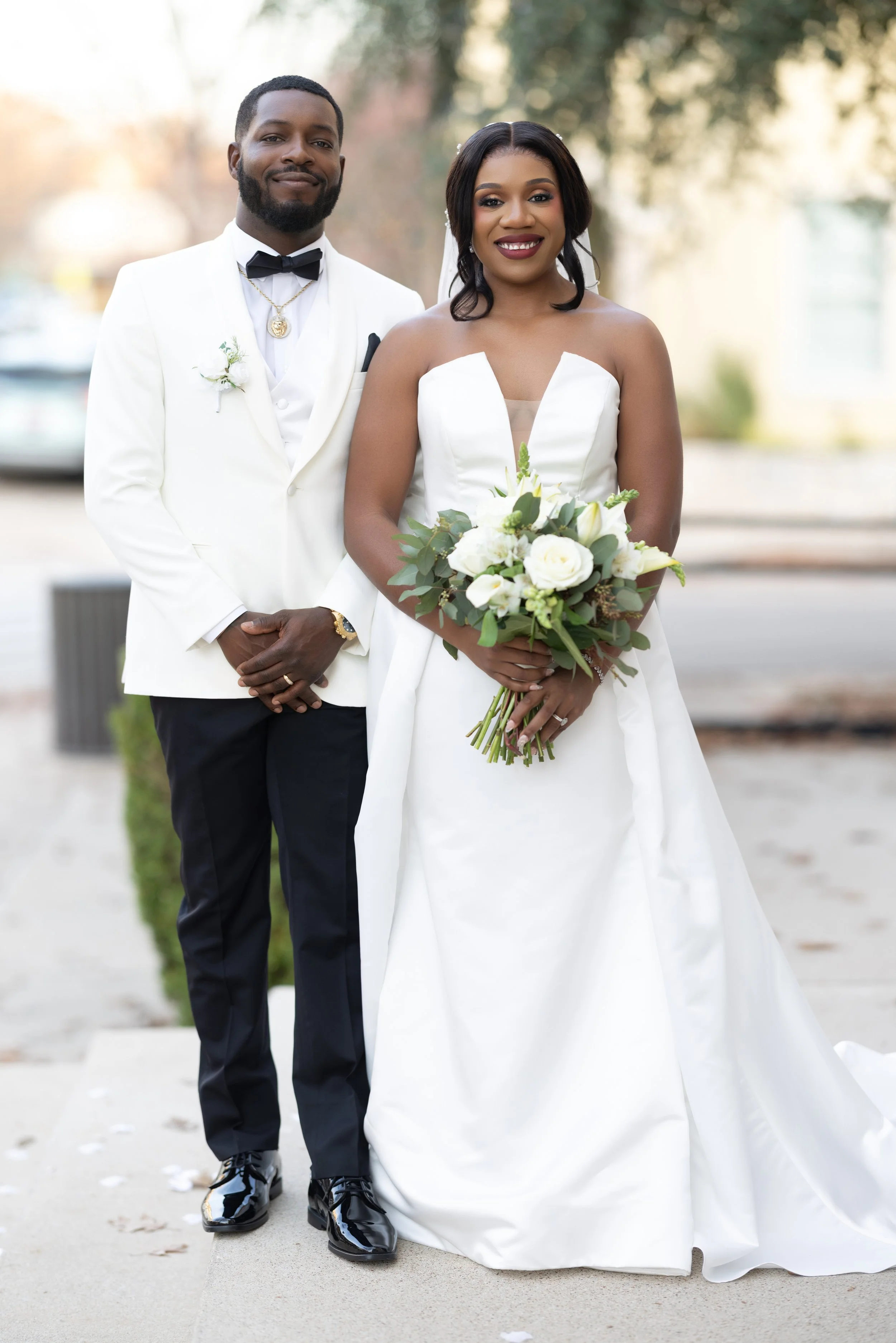 A bride and groom standing outdoors for a wedding photo, with the bride holding a bouquet of white flowers and greenery, both dressed in formal wedding attire.
