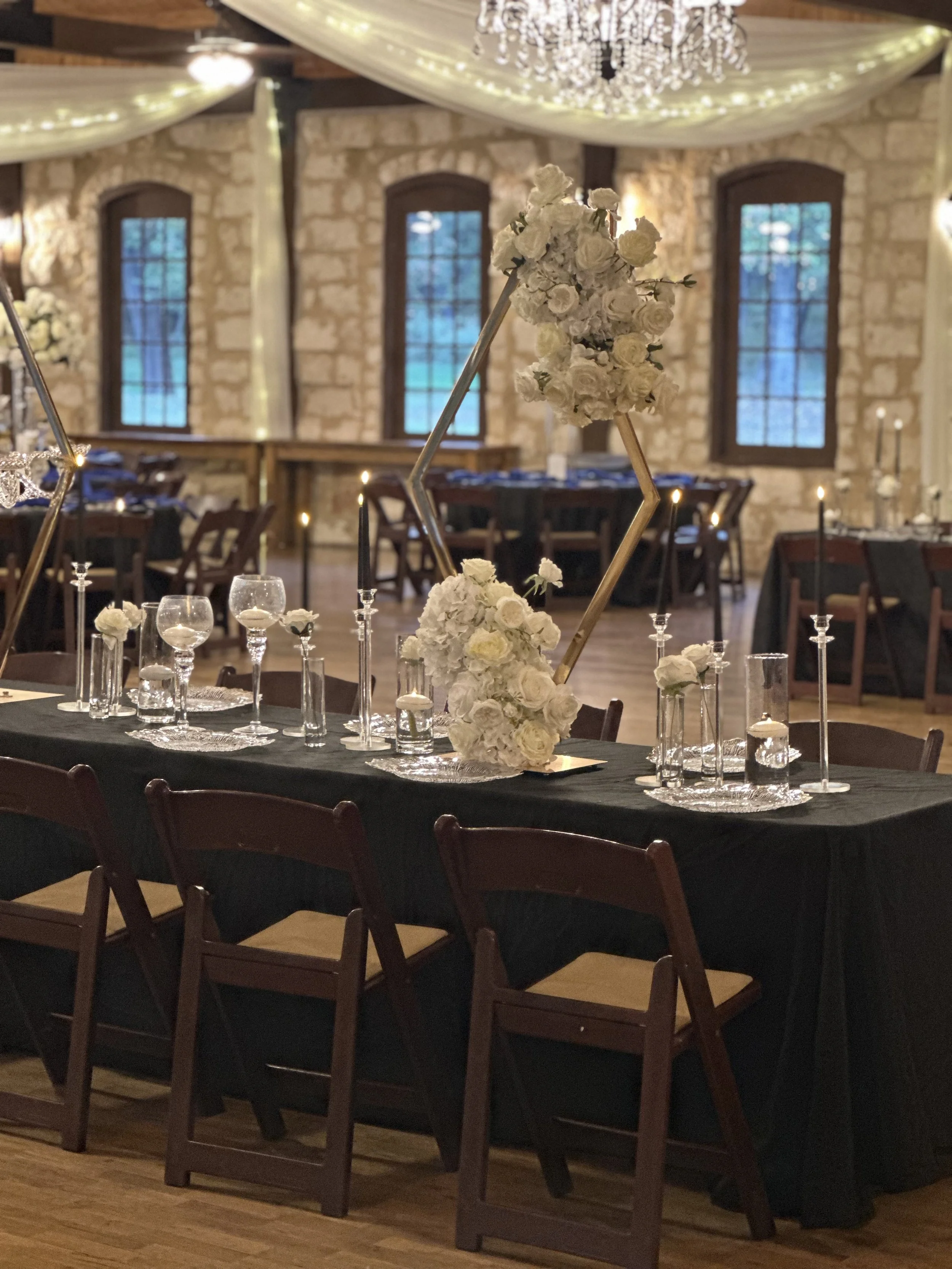 Elegant wedding reception table with white floral centerpieces, black tablecloth, candles, and geometric gold decorations, set in a rustic stone-walled venue with large windows and chandelier.