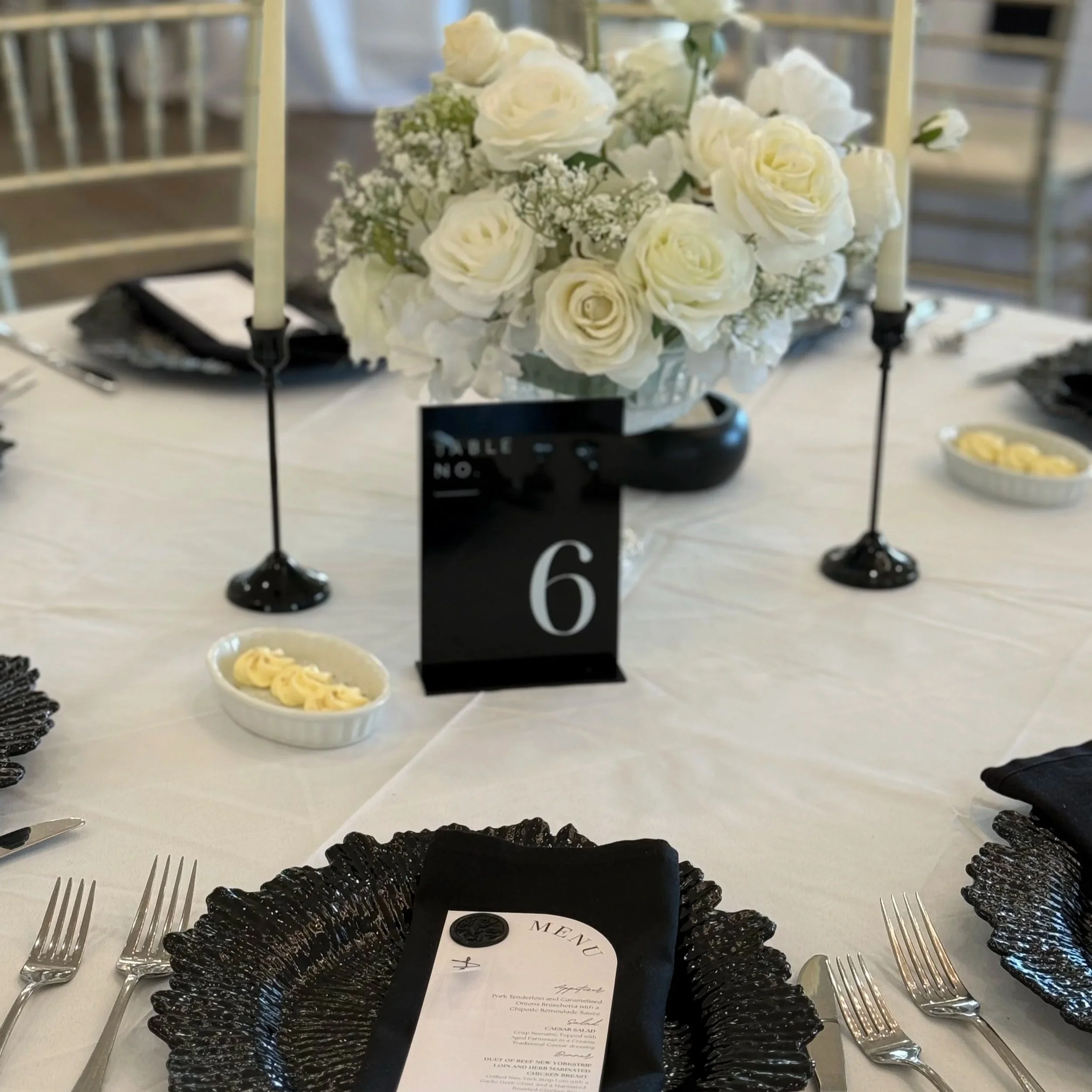 Elegant table setting with a floral centerpiece of white roses, black candle holders, and a black table number sign, with black and white place settings.