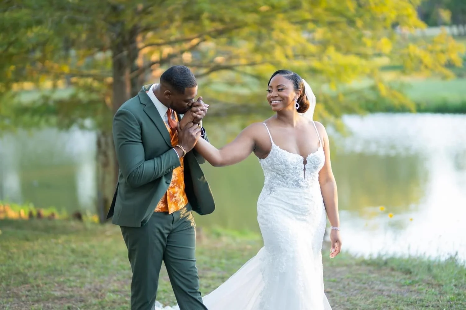 A bride in a white wedding dress smiling as a groom in a suit kisses her hand in a scenic outdoor setting near a lake with trees.
