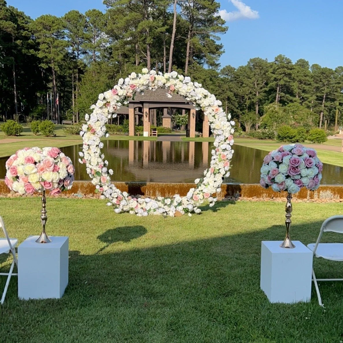 Wedding ceremony setup with a circular floral arch, two flower arrangements on white pedestals, chairs, and a pond in the background, surrounded by greenery and trees.