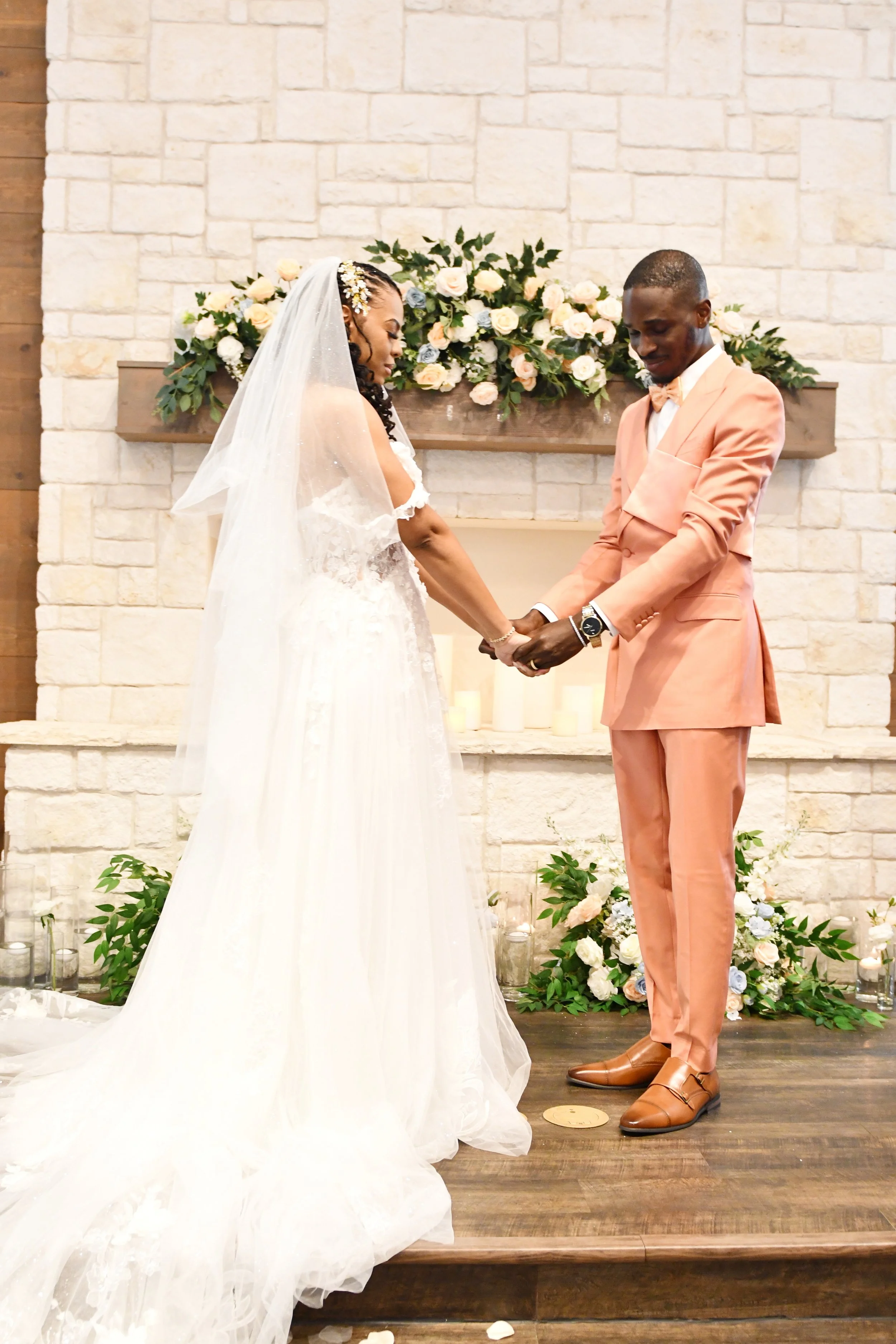 A bride and groom holding hands during their wedding ceremony in front of a decorated fireplace with flowers and candles.