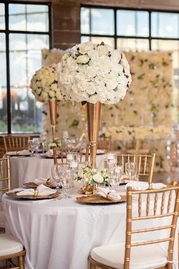 Wedding reception table with tall floral centerpieces of white roses, set with glassware, plates, and gold chiavari chairs, in a rustic venue with large windows.