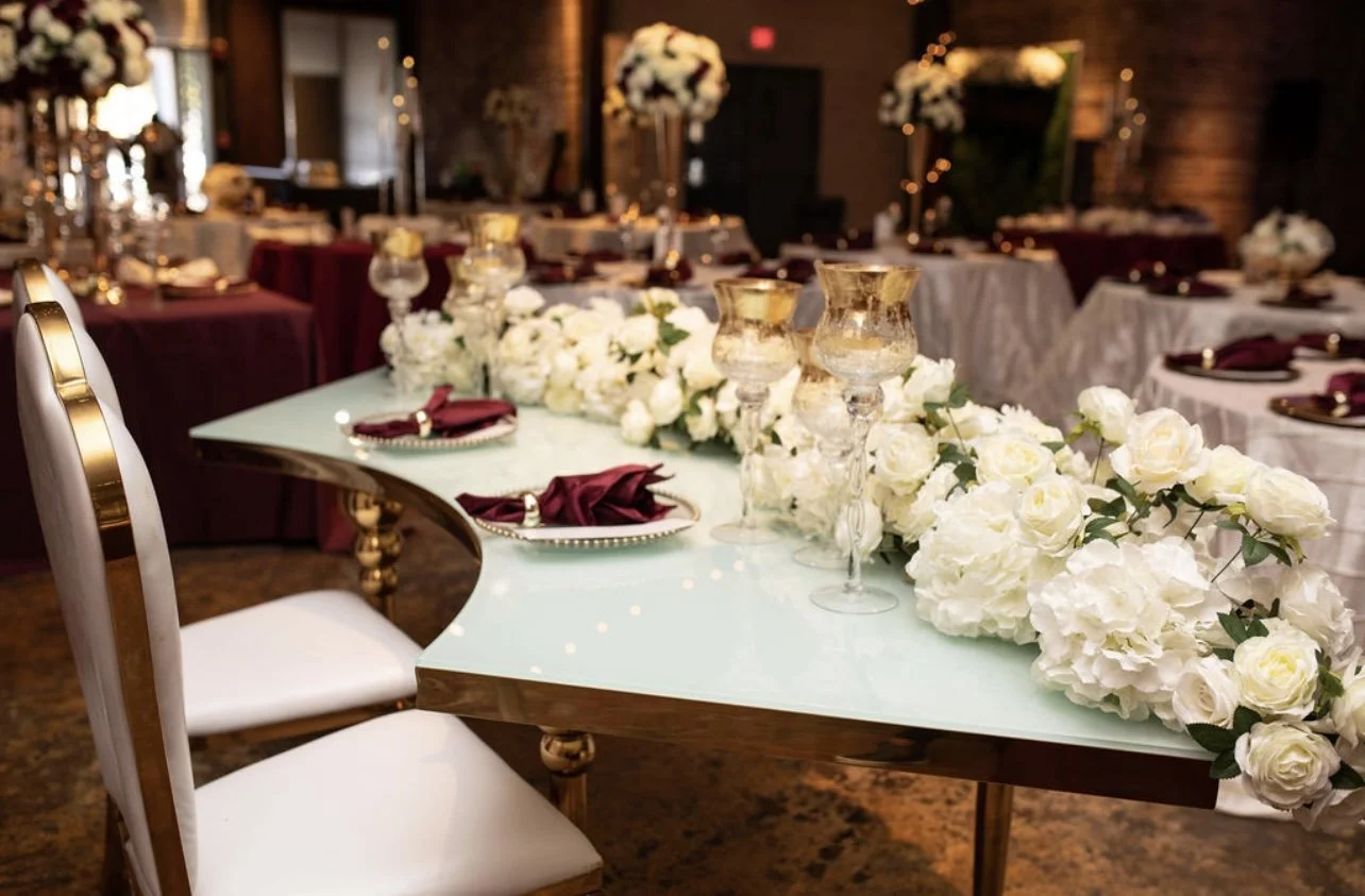 A decorated table with white flowers, gold candles, and burgundy napkins in a dimly lit event space.
