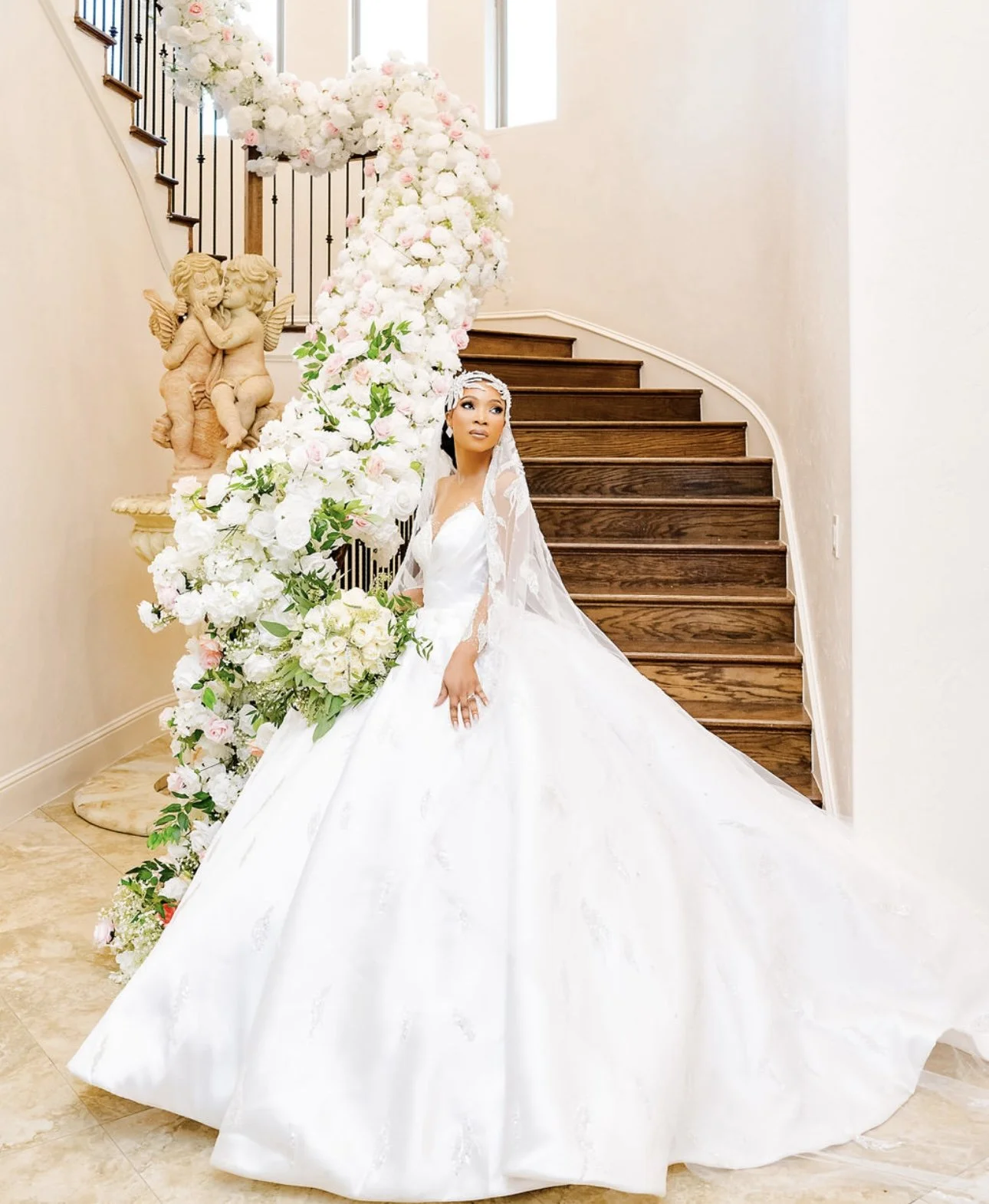 A bride in a white wedding gown and veil standing next to a large floral arch with white and pink flowers, near wooden stairs and a stone cherub sculpture.