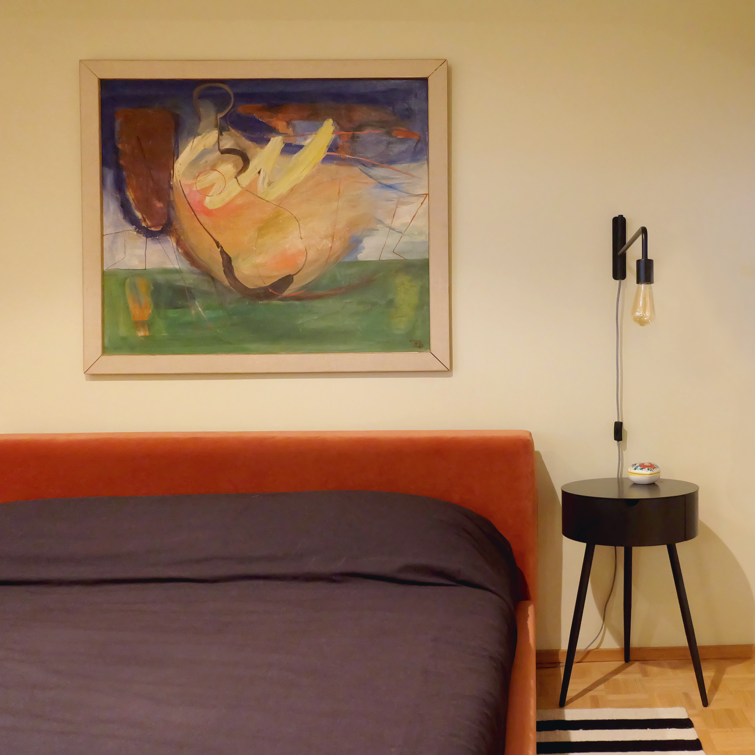 A bedroom corner with a bed, a black side table, a wall-mounted lamp, a small decorative object, a colorful abstract painting on the wall, and a striped rug on the wooden floor.