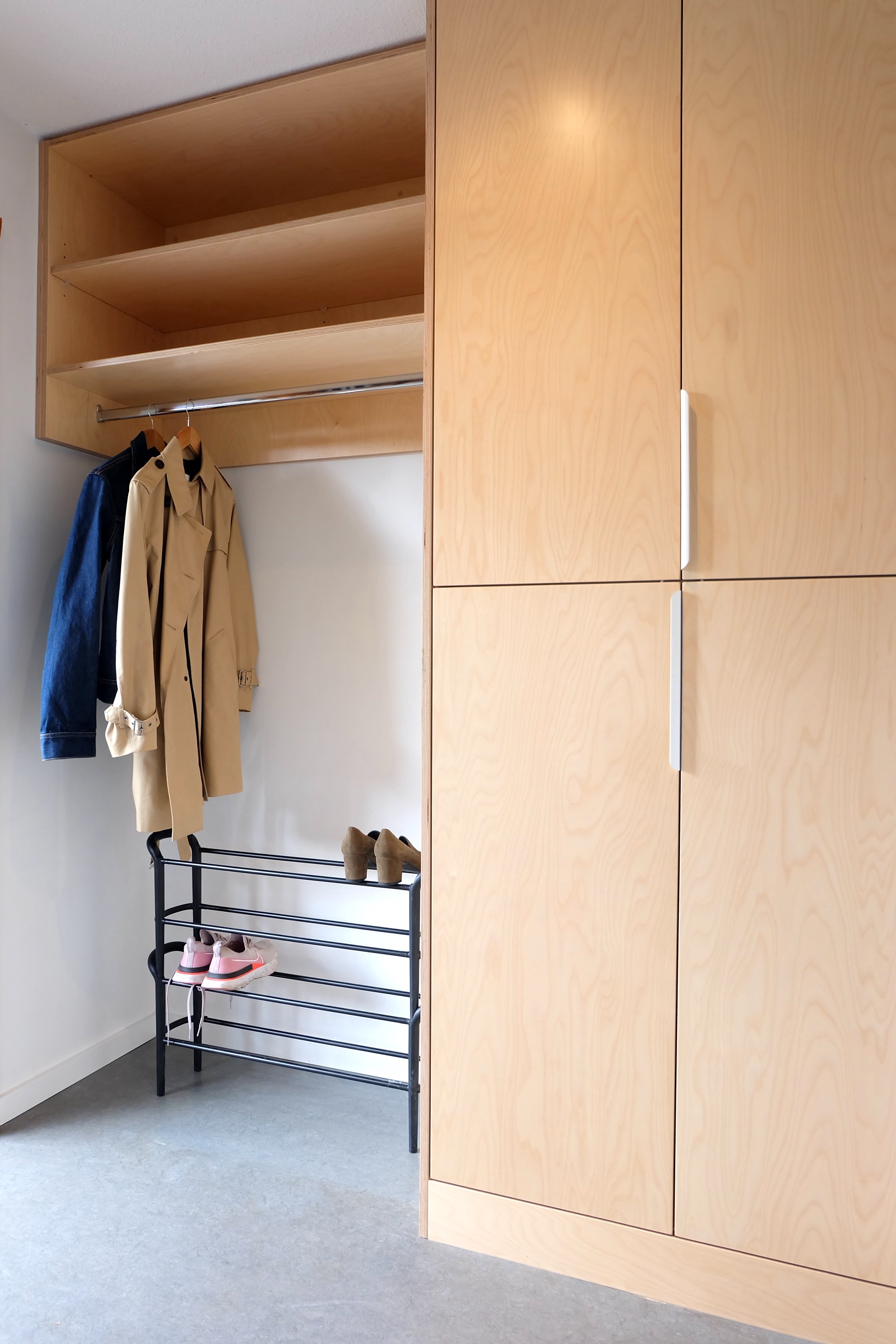 A closet with wooden shelves and a hanging rod holding a beige coat and a denim jacket, with a shoe rack below containing a pair of beige high-heeled shoes and pink sneakers.