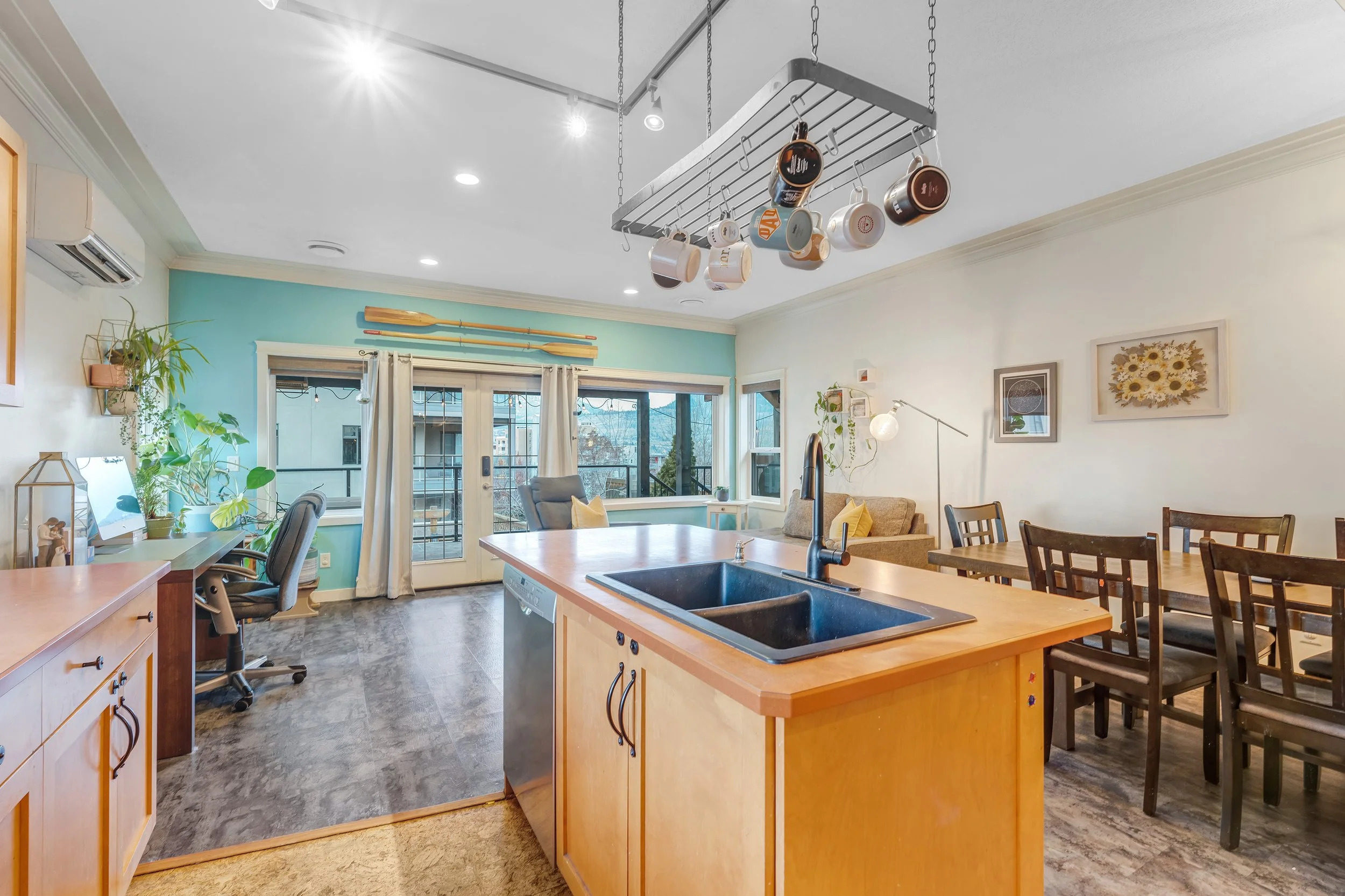 Open-concept living space with a kitchen island, a dining area with a wooden table and chairs, a sitting area with a sofa, large windows with curtains, potted plants, and a balcony door.