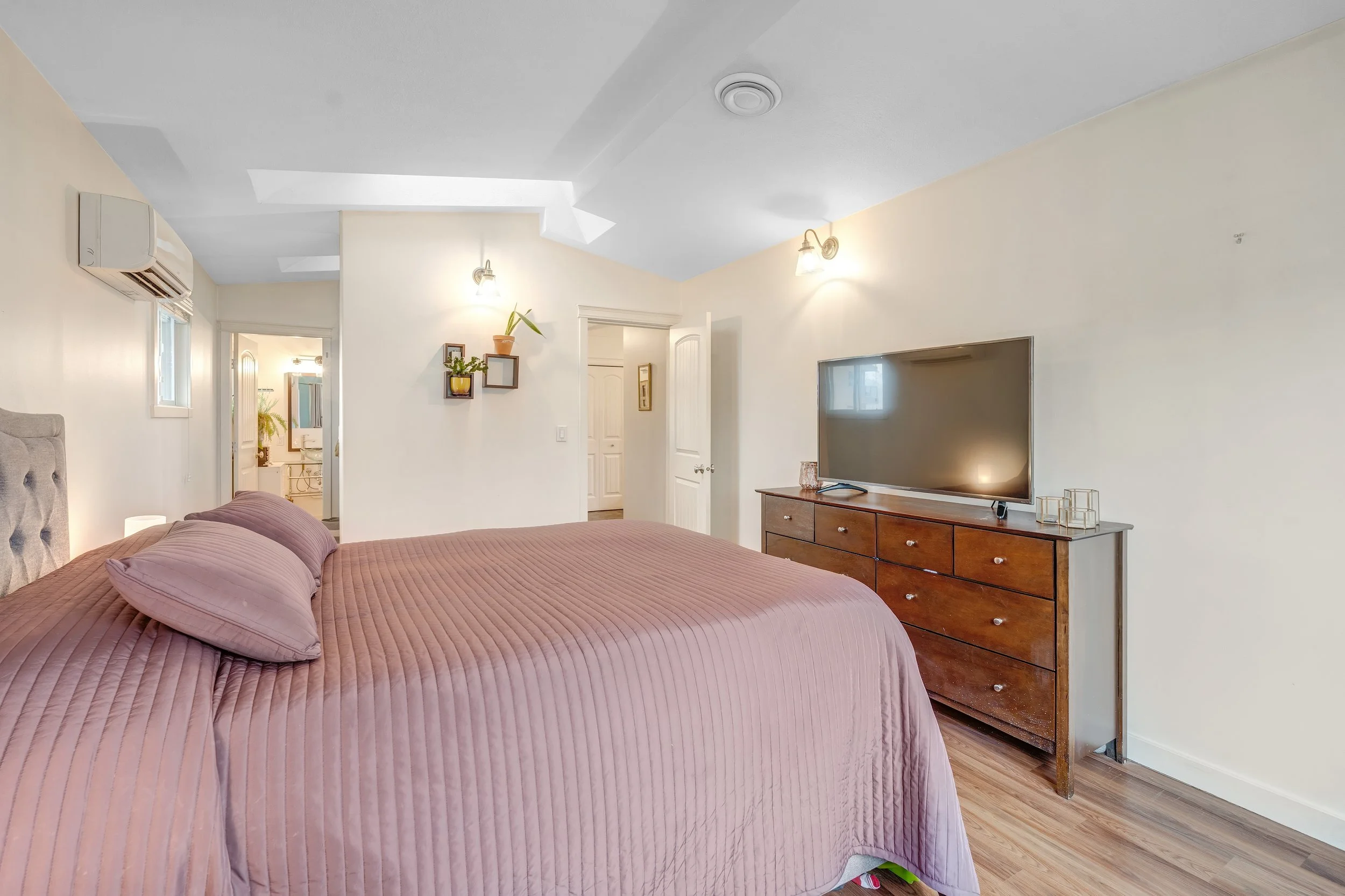 A bedroom with a bed covered in a pink quilt and matching pillows, a wooden dresser with a large flat-screen TV, a small wall shelf with plants, and an air conditioning unit on the wall.