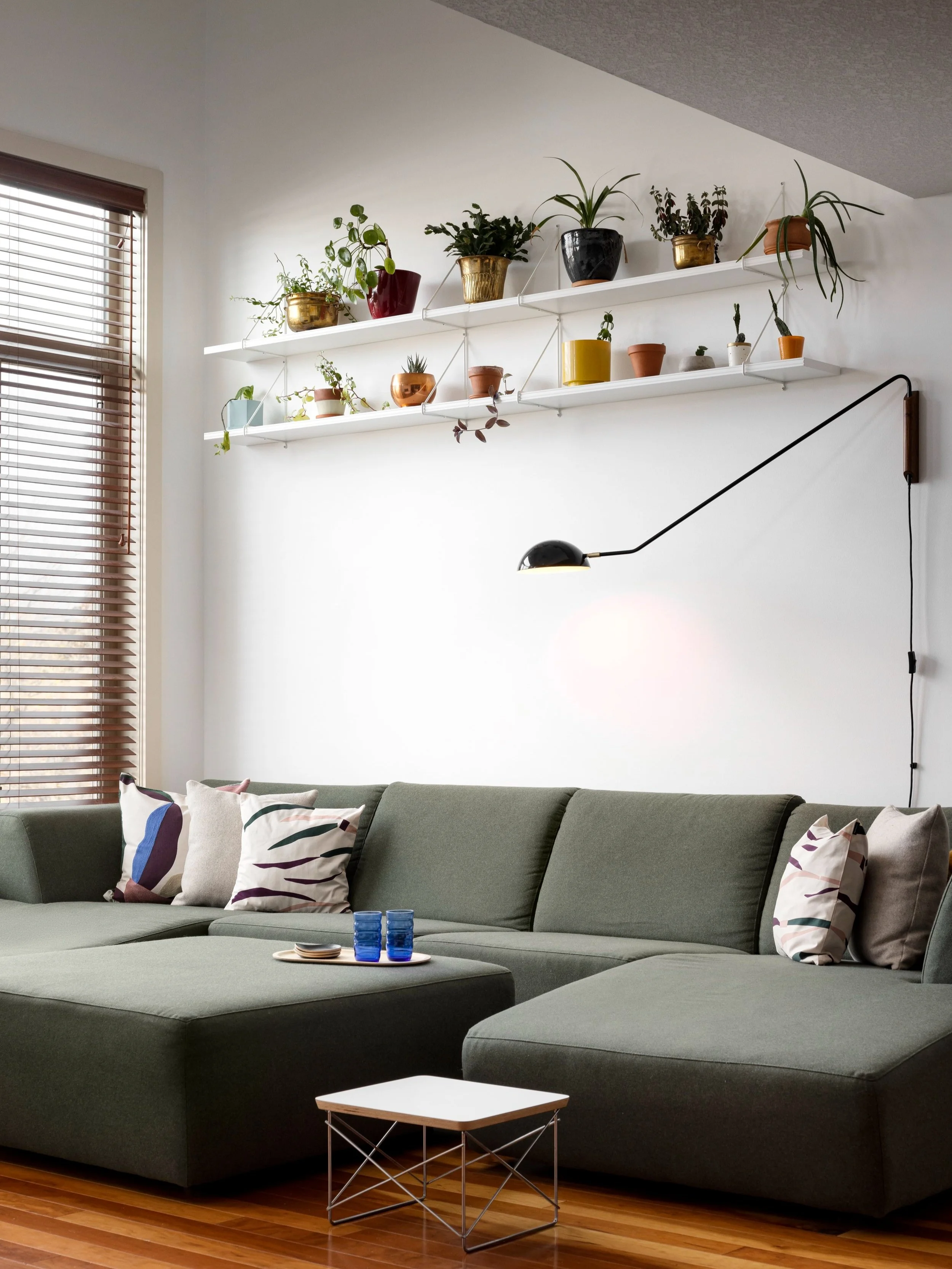 Living room with green sectional sofa, patterned throw pillows, a small white and metal side table, and a wall-mounted black lamp. There are two shelves above the sofa holding various potted plants. A large window with wood blinds is on the left.