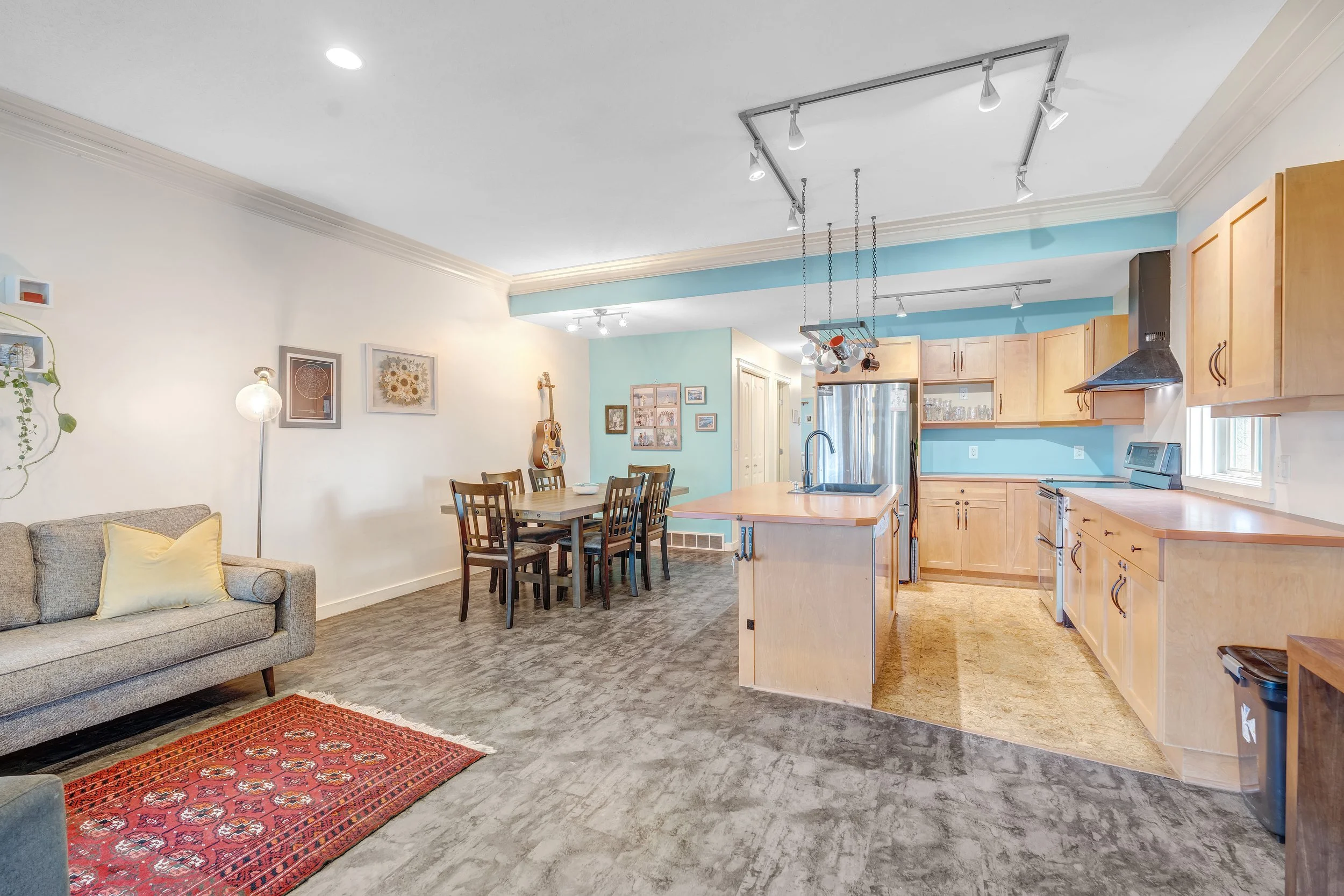 Open-concept living room and kitchen with light-colored cabinetry, stainless steel appliances, a dining table with chairs, a sofa with a yellow pillow, and a red patterned rug.