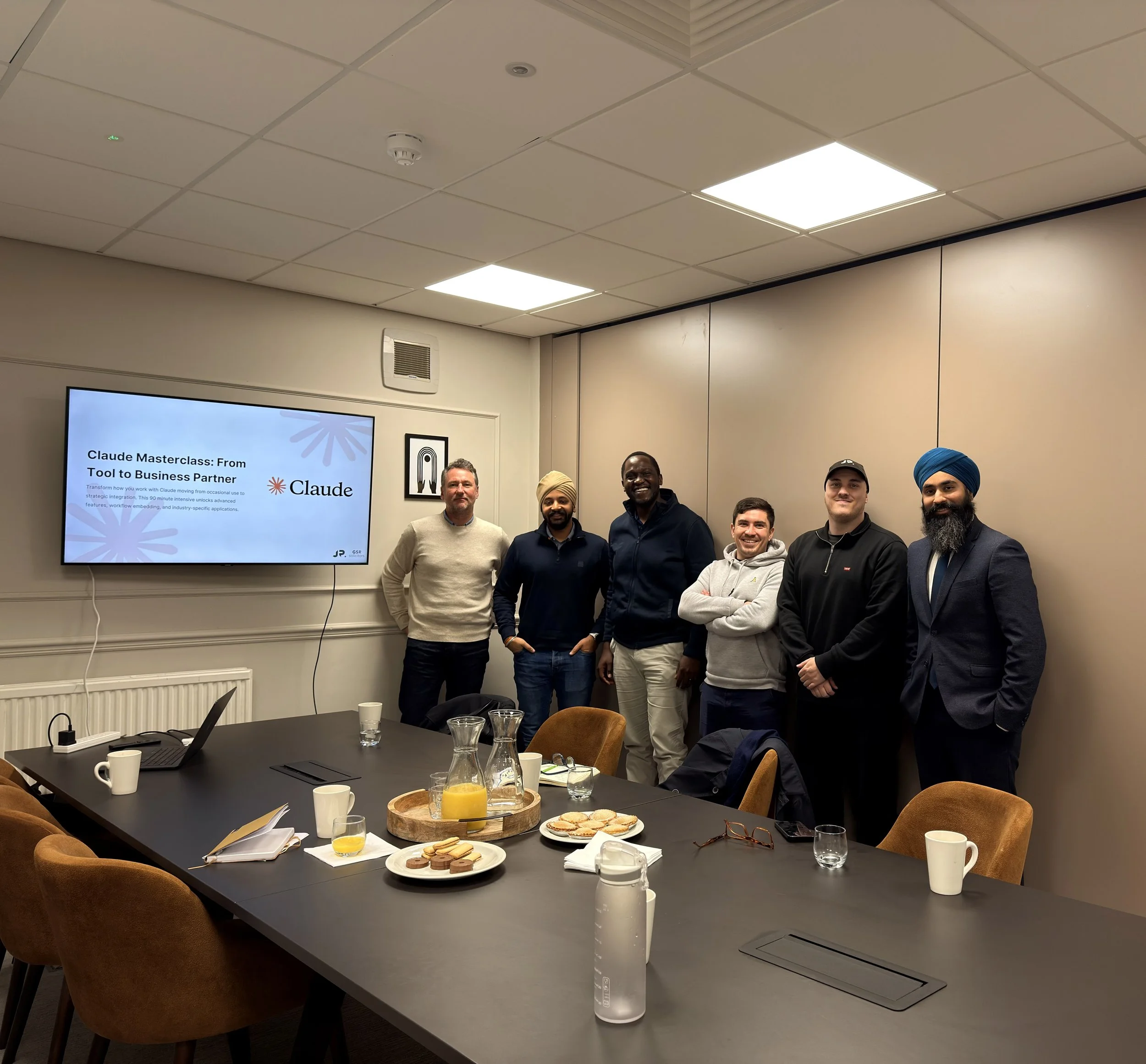Six men standing in a conference room in front of a wall and a large TV screen. The table in front is set with drinks, snacks, and a laptop. The screen displays a presentation slide titled 'Claude Masterclass: From Tool to Business Partner'.