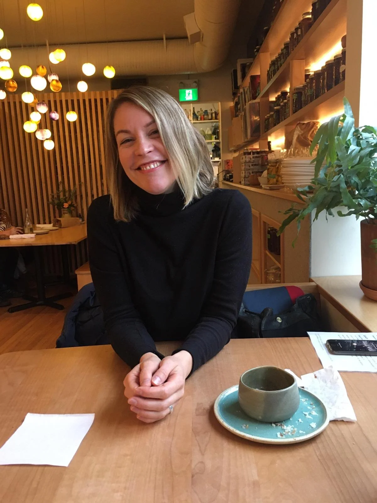 A woman with shoulder-length blonde hair and a black long-sleeve top, smiling and sitting at a wooden table in a cozy, well-lit cafe with hanging round lights and a bookshelf in the background. There is a green teacup and saucer with some crumbs on the table.