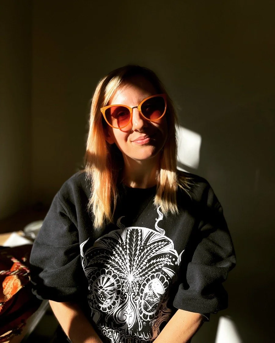 A woman wearing large orange sunglasses and a black sweatshirt with a white detailed skull design, sitting in a room with sunlight casting shadows on her face and background.
