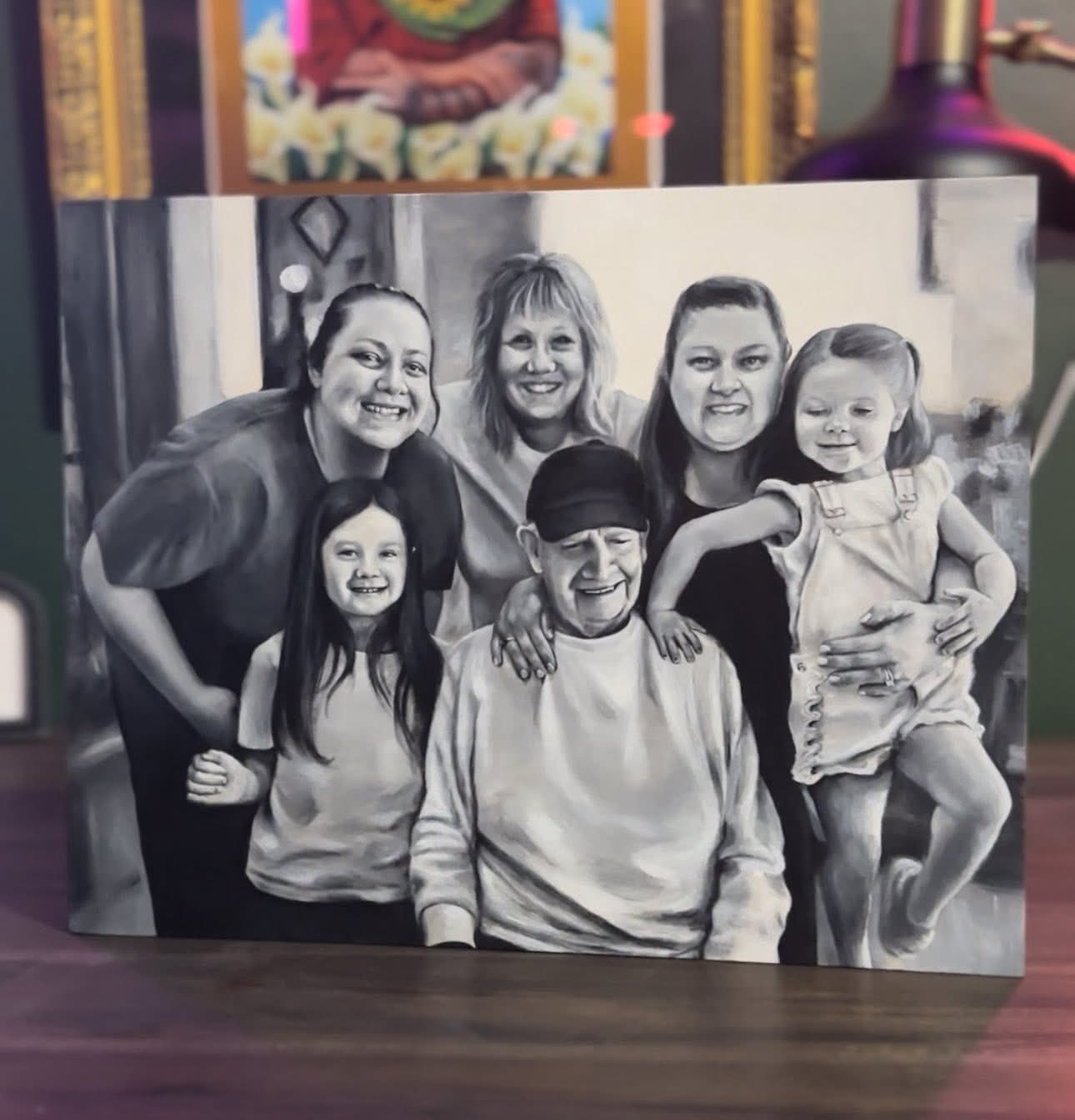 Black and white photo of six people, including children and adults, smiling and posing together, with some leaning on each other, indoors with framed pictures on the wall.