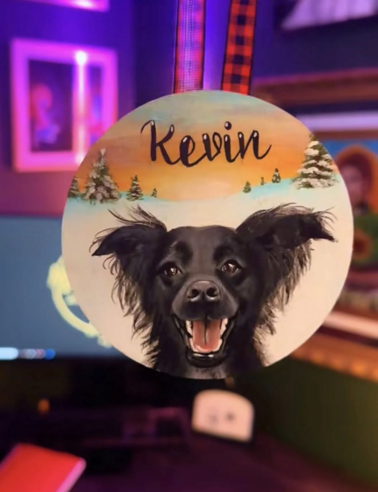 Decorative round ornament featuring a dog's face with large ears and open mouth, against a painted snowy landscape background with trees and a sunset sky, and the name 'Kevin' written at the top.