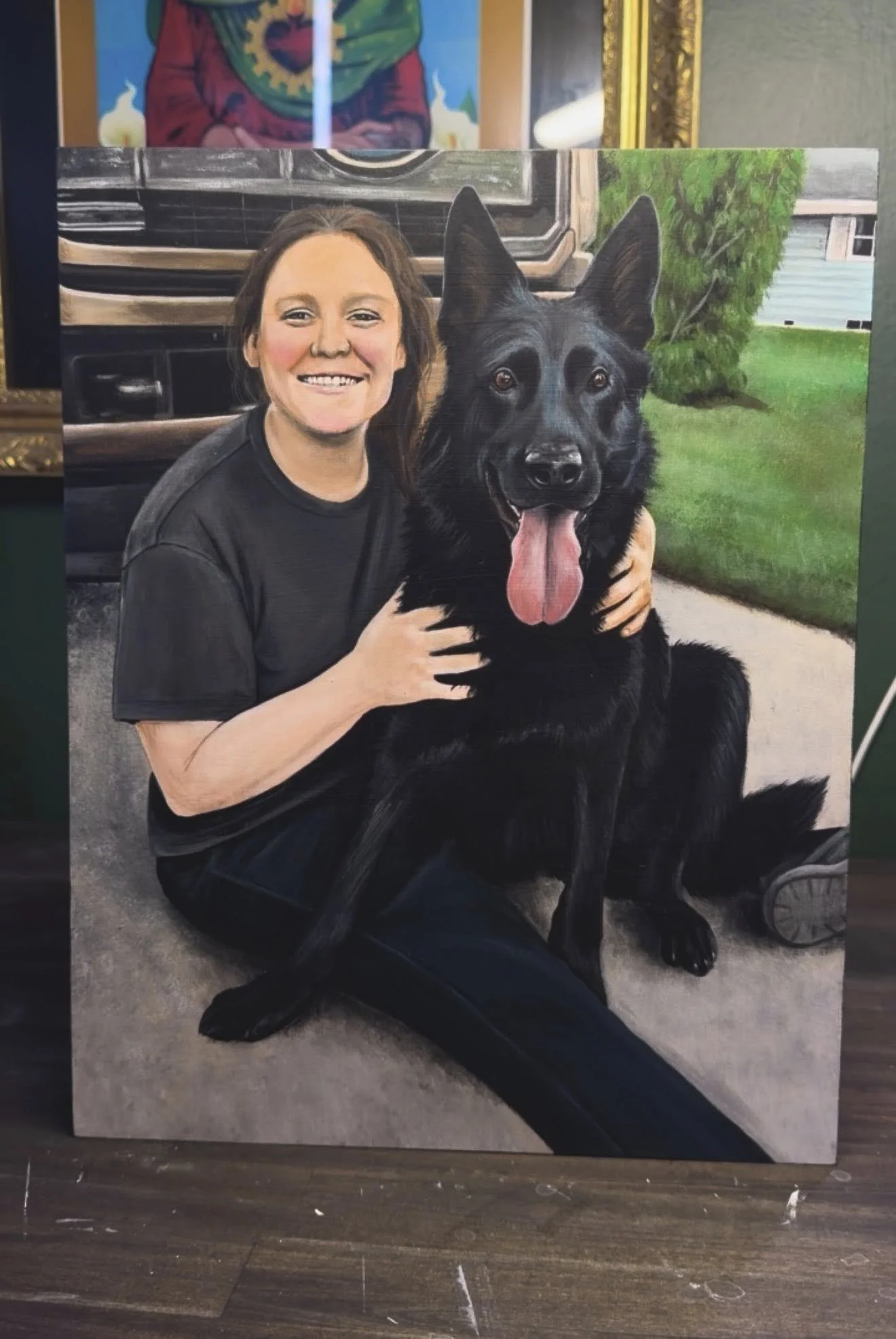 A woman sitting on the floor with a large black German Shepherd dog, smiling, outdoors near a green lawn and a house. The woman is wearing a black T-shirt and the dog has its tongue out.