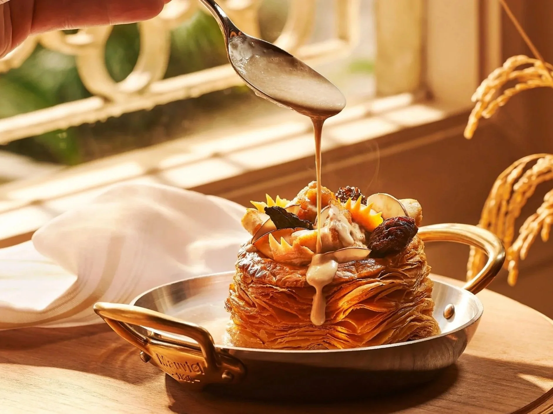 A layered croissant pastry topped with dried fruits and garnishes, with honey being drizzled over it from a spoon.