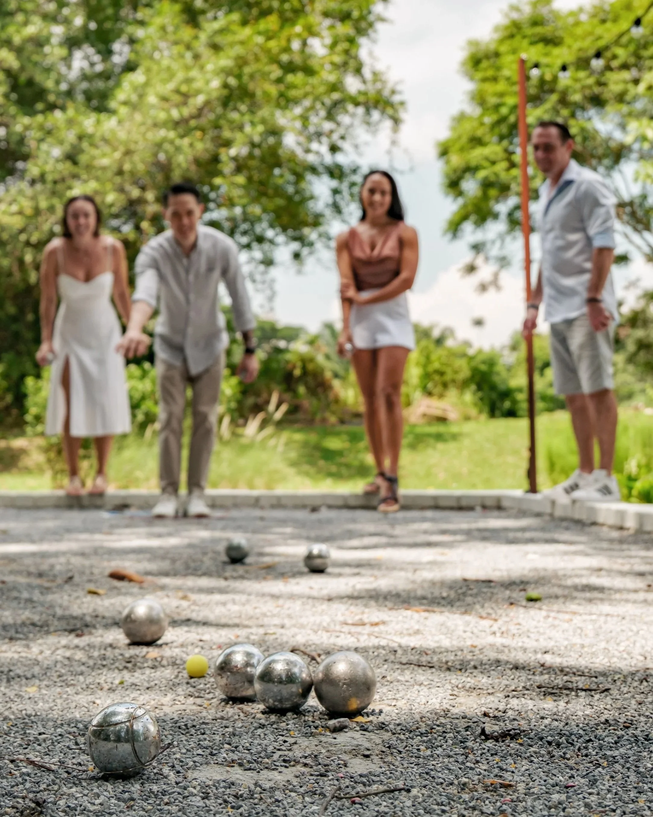 People playing petanque outdoors on a gravel court with trees in the background.