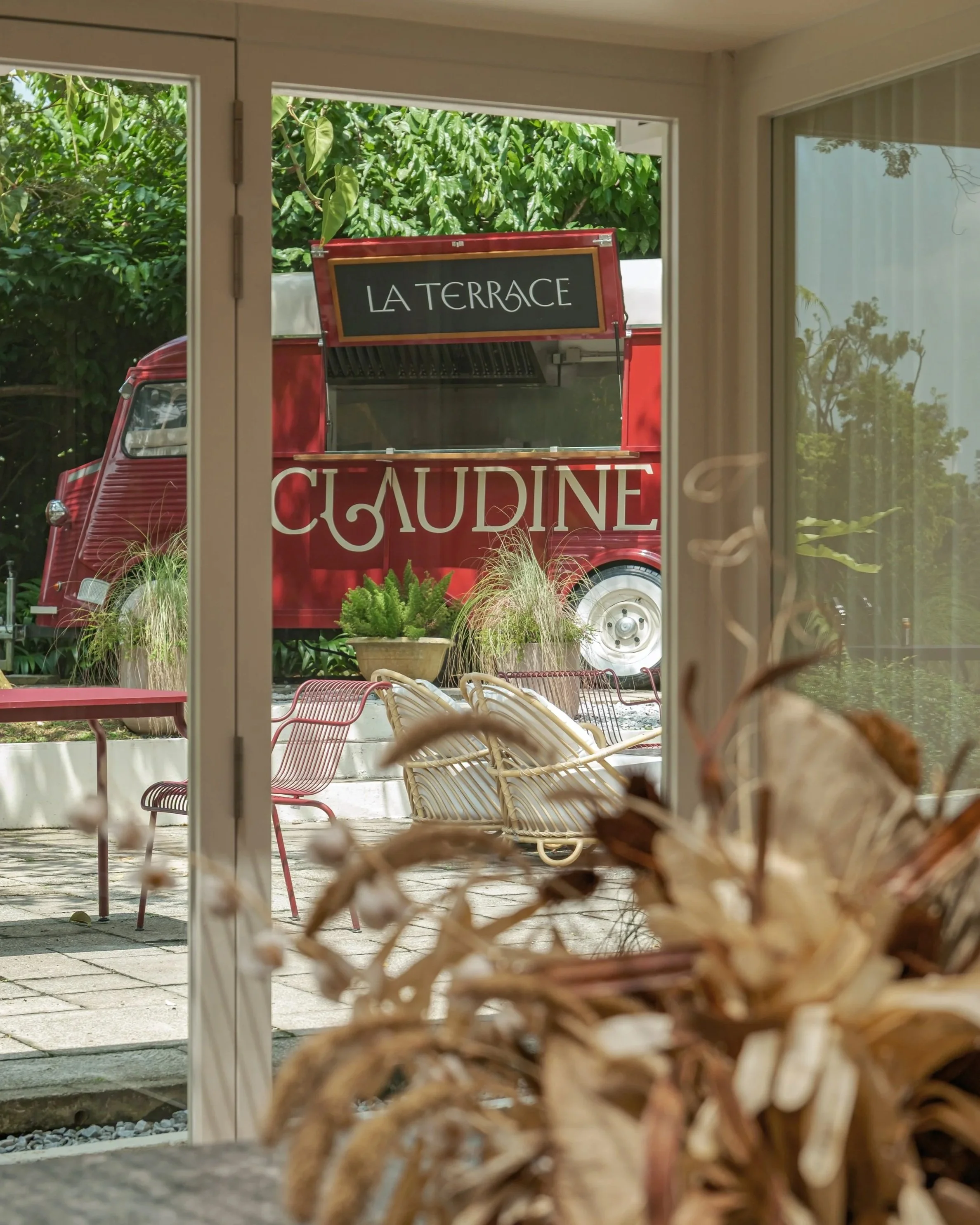 View through a window showing a red food truck with the sign ‘LA TERRACE’ parked outside, surrounded by greenery and outdoor seating.