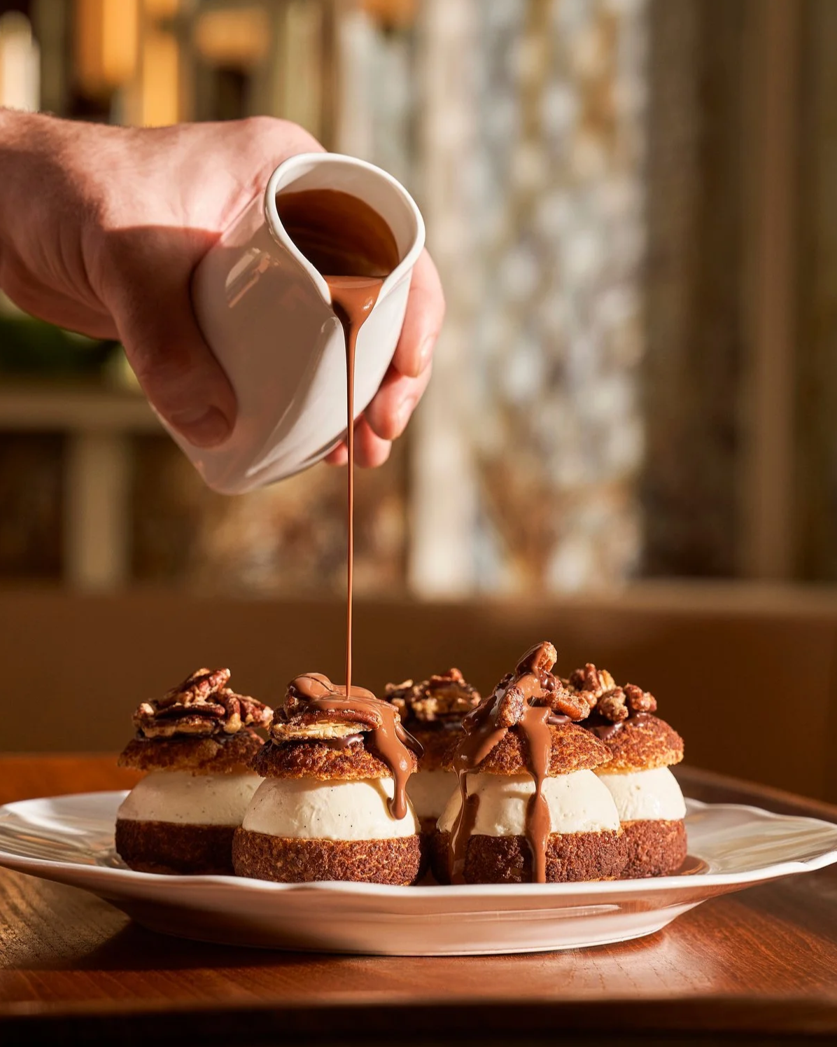 A hand pouring chocolate sauce over dessert pastries on a plate.