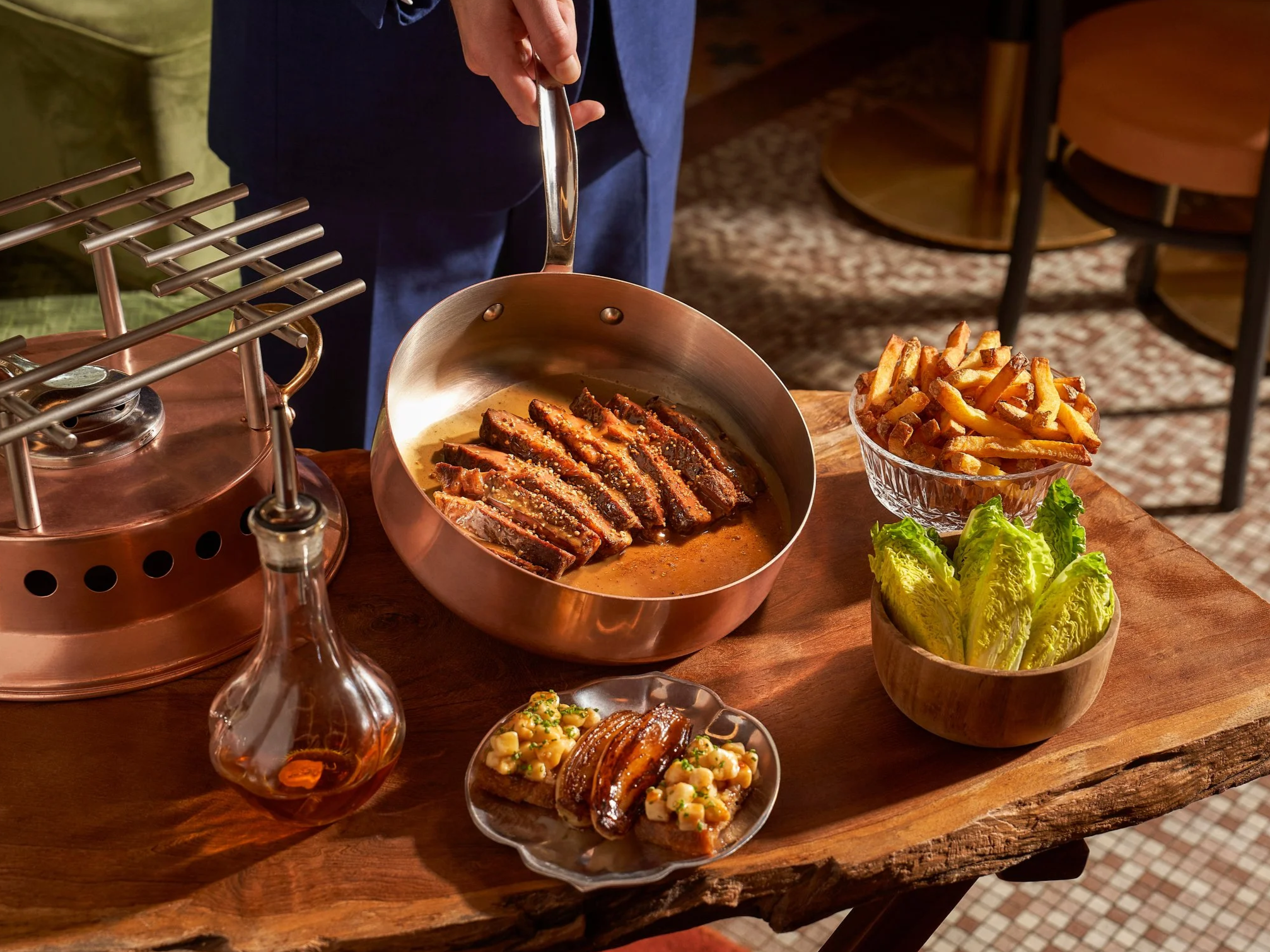 A person cooking sliced steak in a copper pan, surrounded by a bowl of French fries, a bowl of lettuce, a small dish of food, and a bottle of sauce on a rustic wooden table.