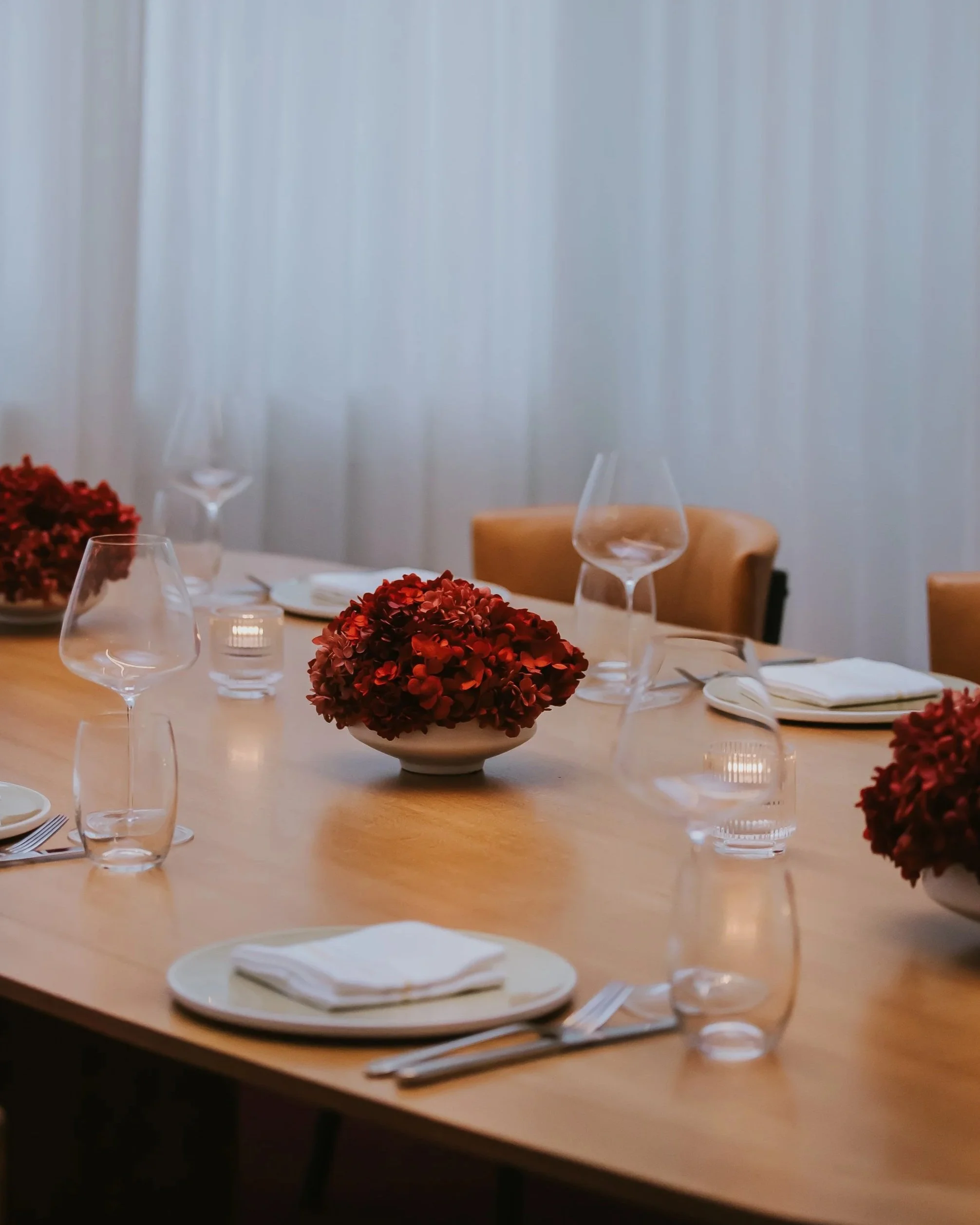 A dining table set with plates, napkins, glasses, forks, and knives, decorated with red floral centerpieces.