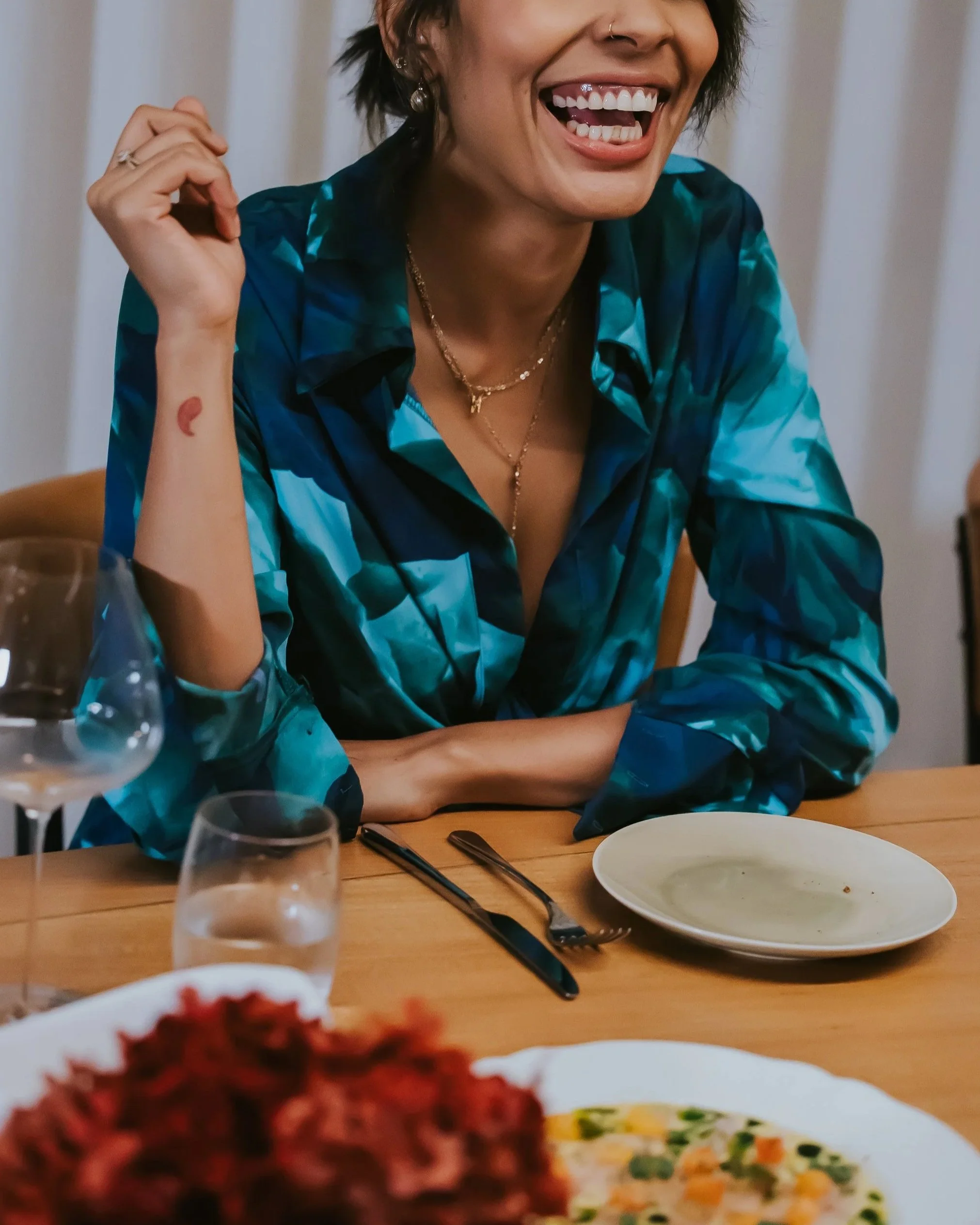 A woman laughing at a dinner table, wearing a blue patterned shirt, gold jewelry, and a nose ring, with food and drinks on the table.