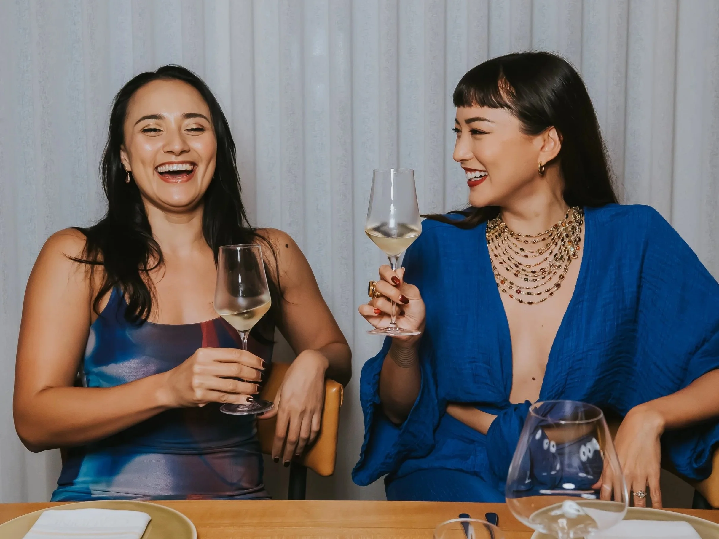 Two women in blue dresses sitting at a dinner table, laughing and holding glasses of white wine, in front of a white curtain.