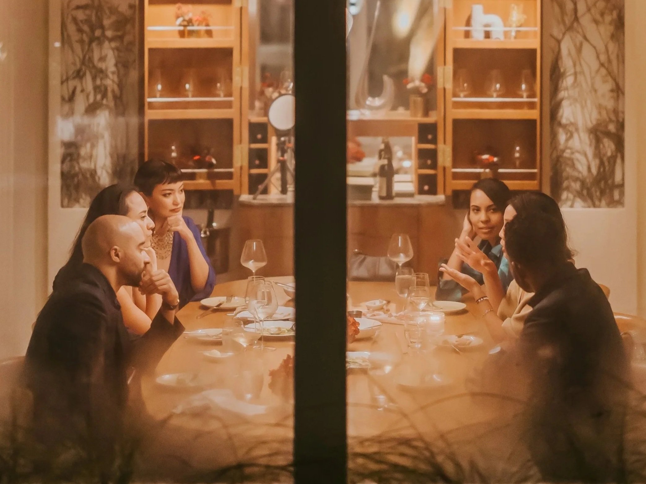 People seated at a dining table having a conversation in a warmly lit restaurant, with a wooden shelf in the background holding bottles and glasses.