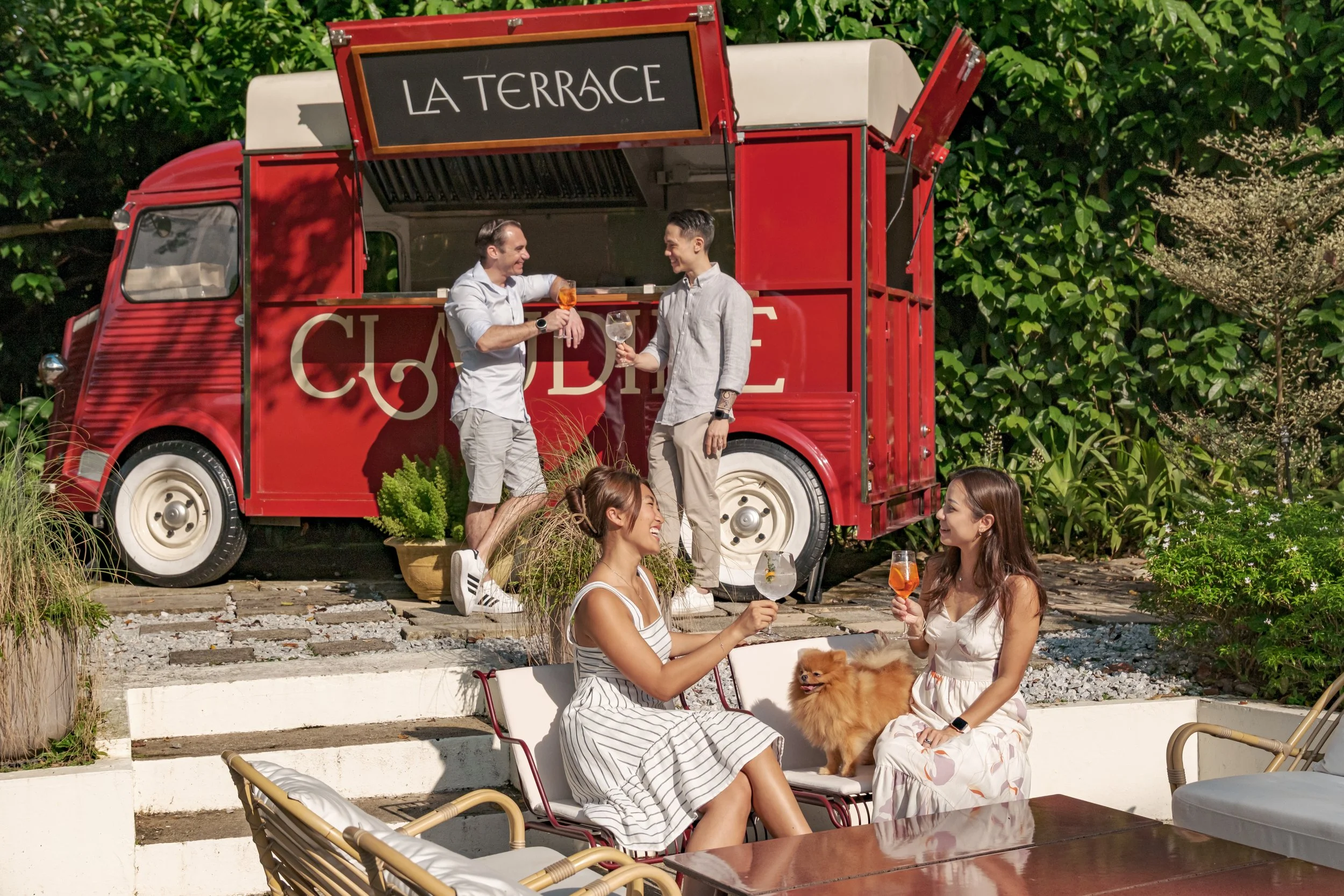 People enjoying drinks and conversation outdoors in front of a vintage red food truck with a sign that reads 'La Terrasse'. Two women are sitting on chairs, one dog with fluffy fur sitting on one woman's lap, and two men are standing on the food truc