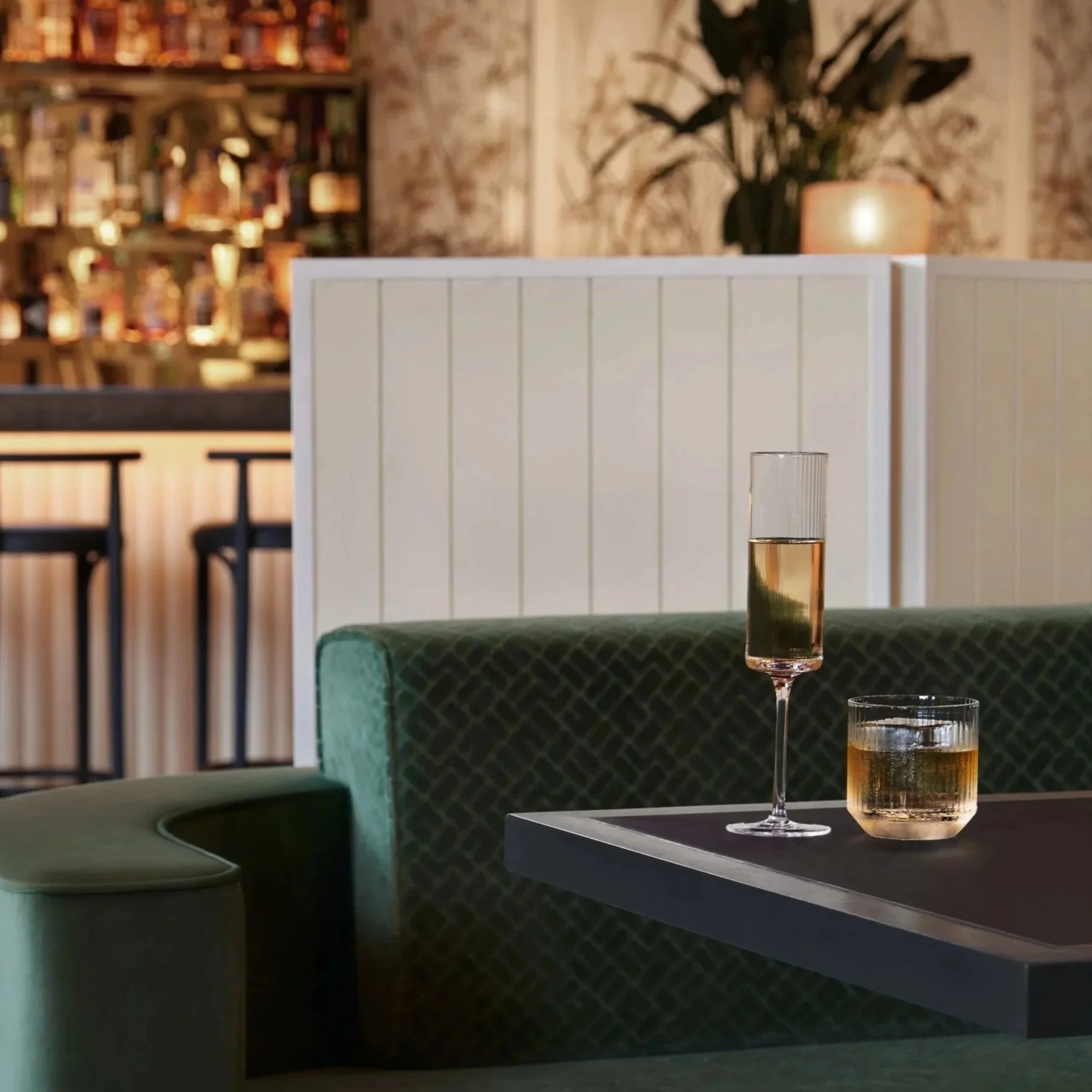 A bar or lounge with a green upholstered couch and a black table holding two glasses of drinks, one with champagne and the other with whiskey. The background features a white partition, a bar counter, and a potted plant with blurred shelves.