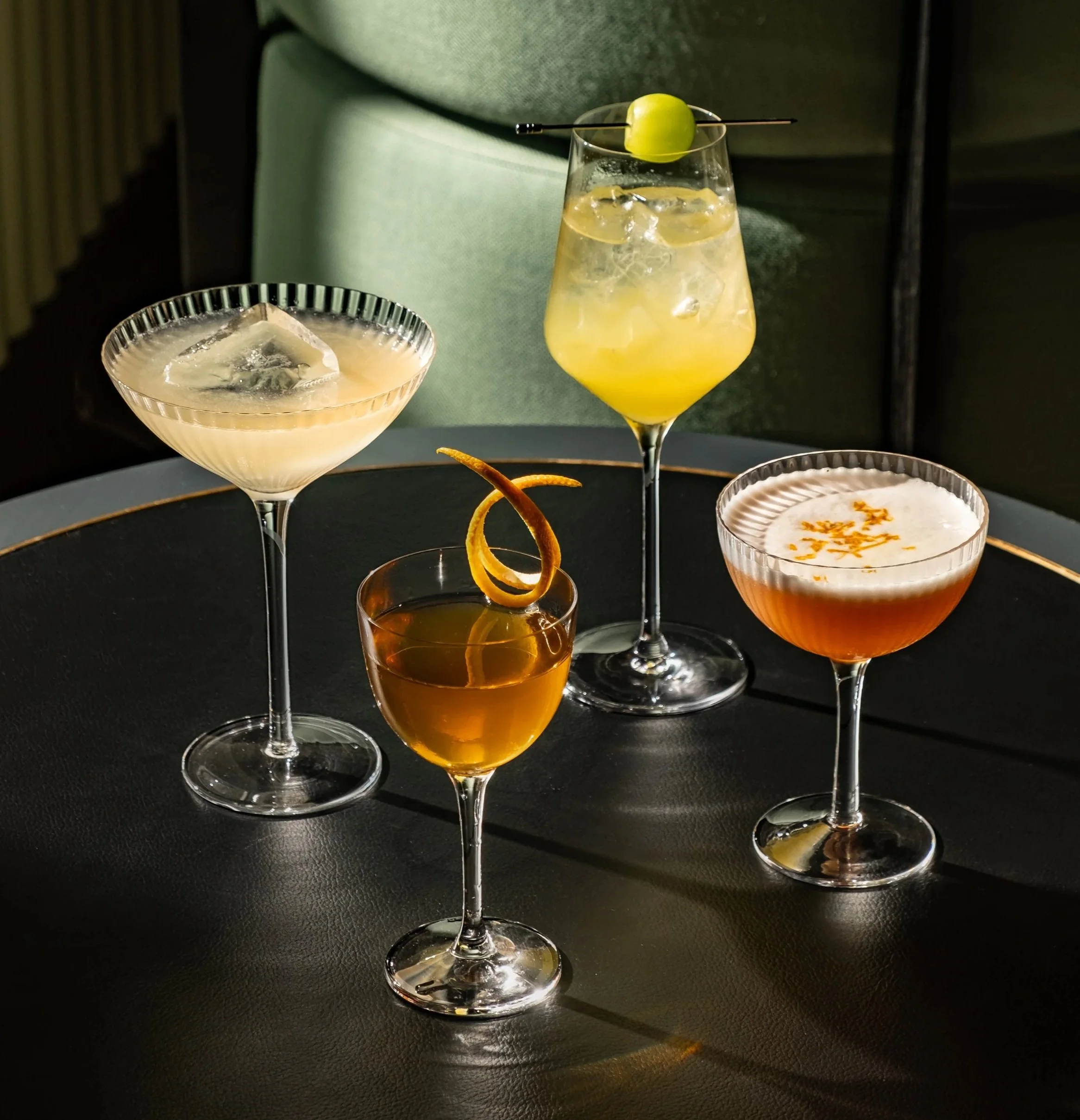 Four different cocktails on a black table, each in a unique glass with garnishes, with a green upholstered seat in the background.