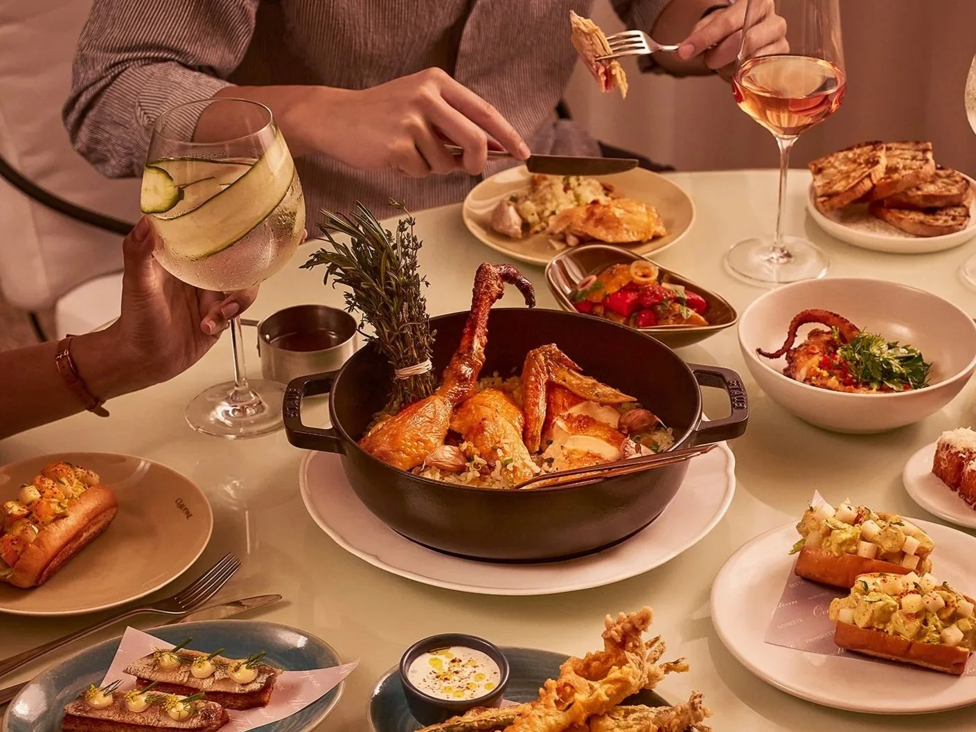 People enjoying a dinner with various dishes including roasted chicken, salads, bread, and drinks, including wine and a cocktail with cucumber slices.
