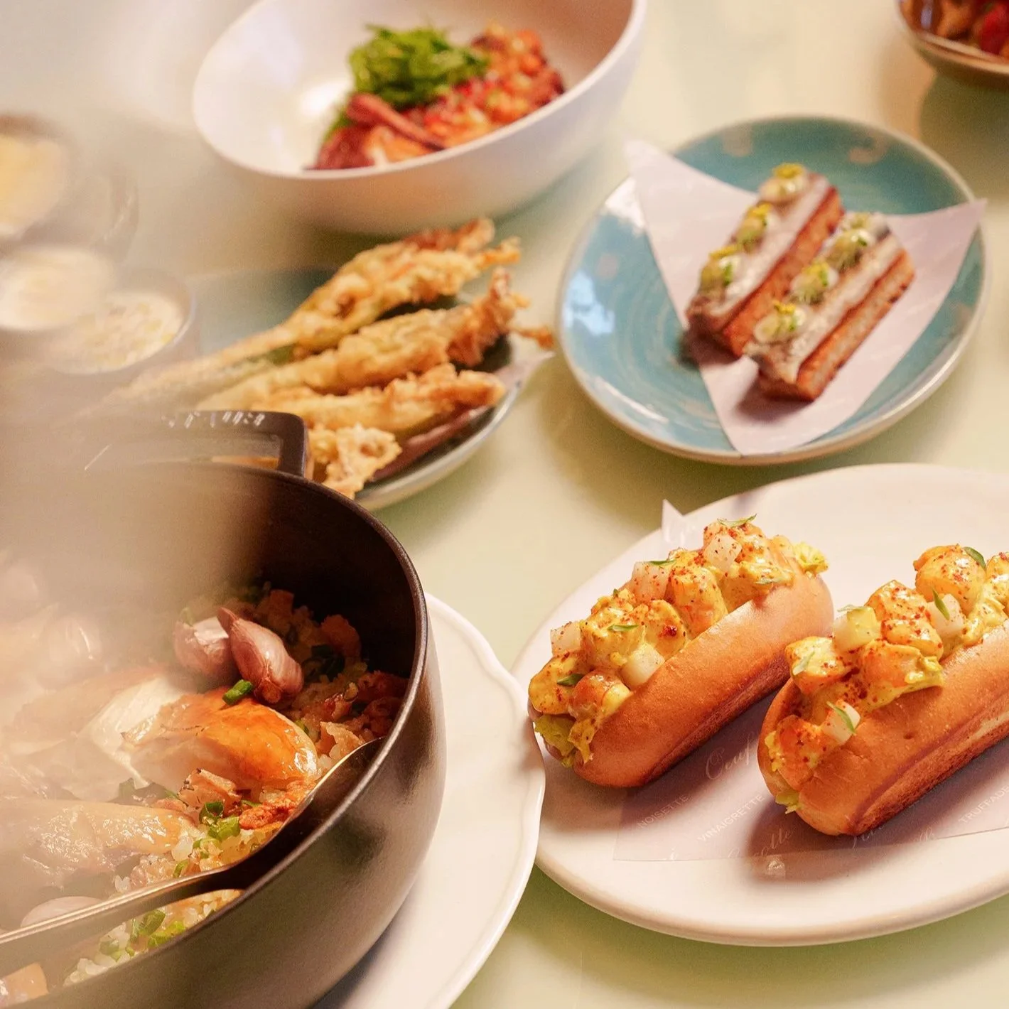 A variety of Asian dishes including sushi, tempura, and hot dogs with seafood topping, served on different plates and bowls.