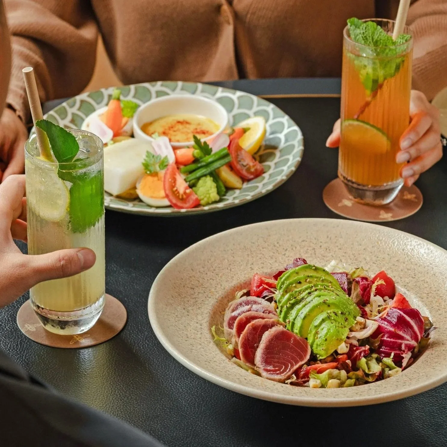 Two people holding glasses of lemonade with mint and lemon slices, a plate of salad with sliced avocado, tomatoes, and red onions, and another plate of tuna salad with avocado and mixed greens on a black table.