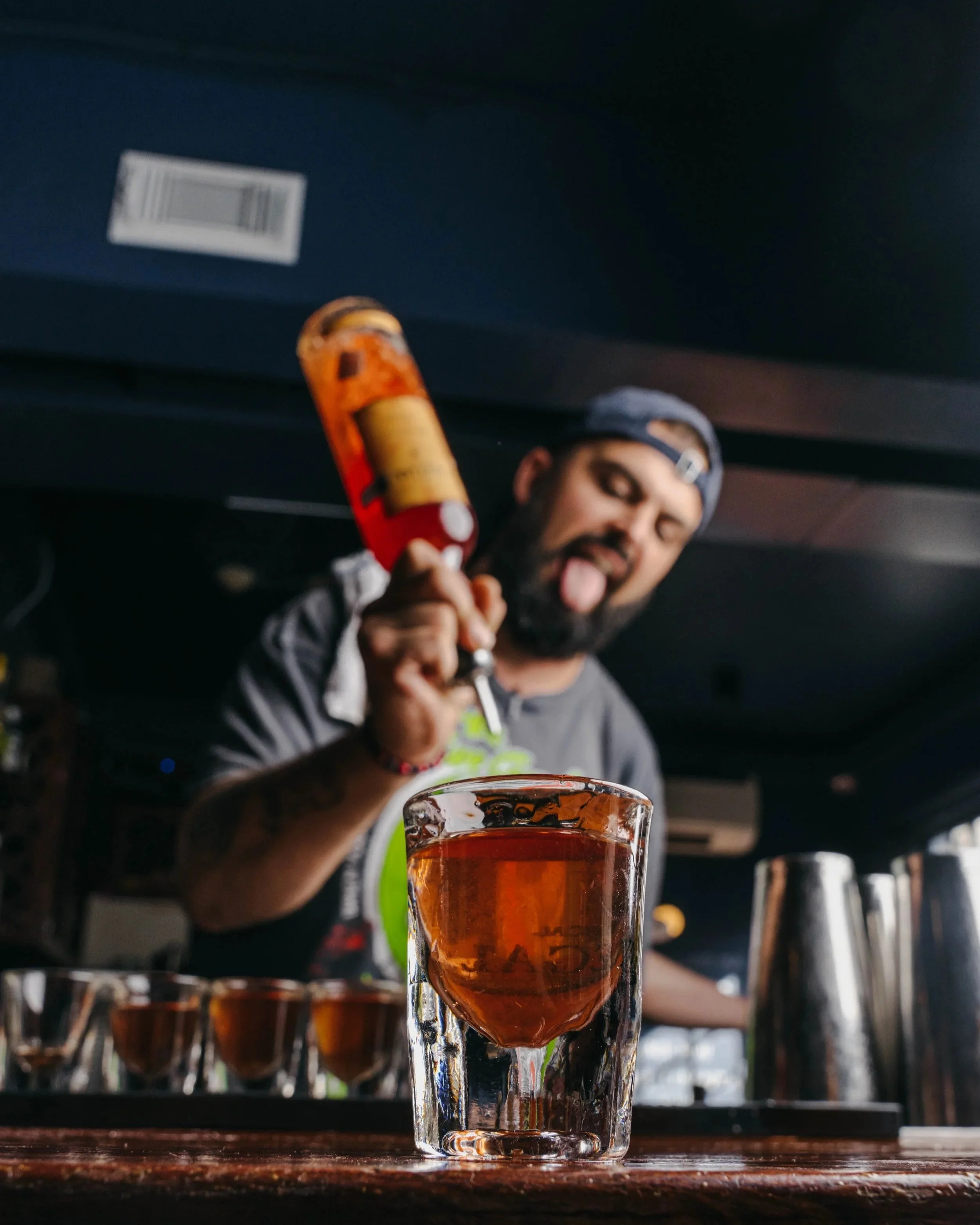 A bartender holding a bottle of liquor behind a glass of amber-colored drink on a bar counter, with bar tools and small glasses in the background, and the bartender sticking out his tongue.
