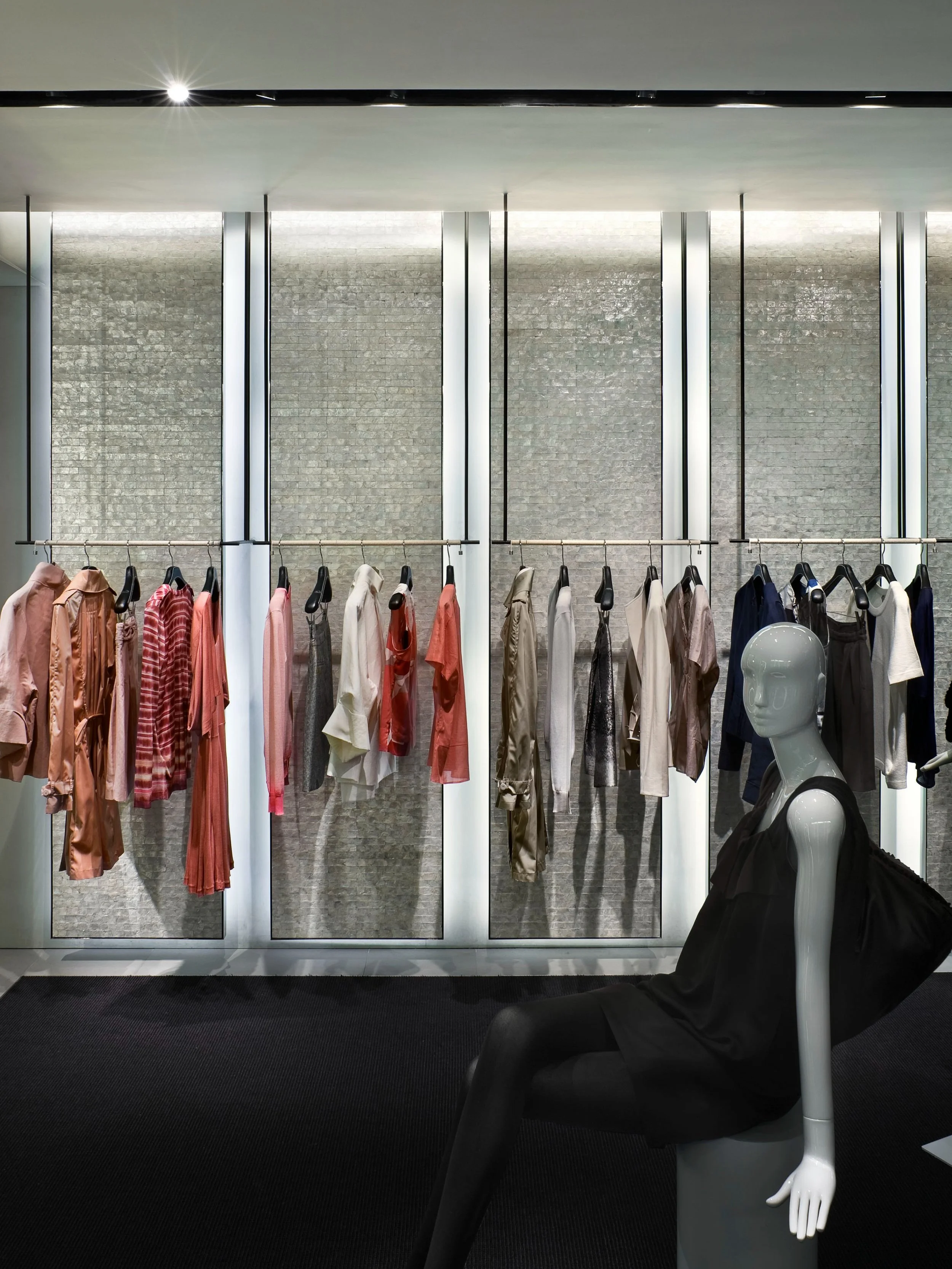 JGNA retail interior with garment rails against a textured mosaic wall and a seated mannequin in black