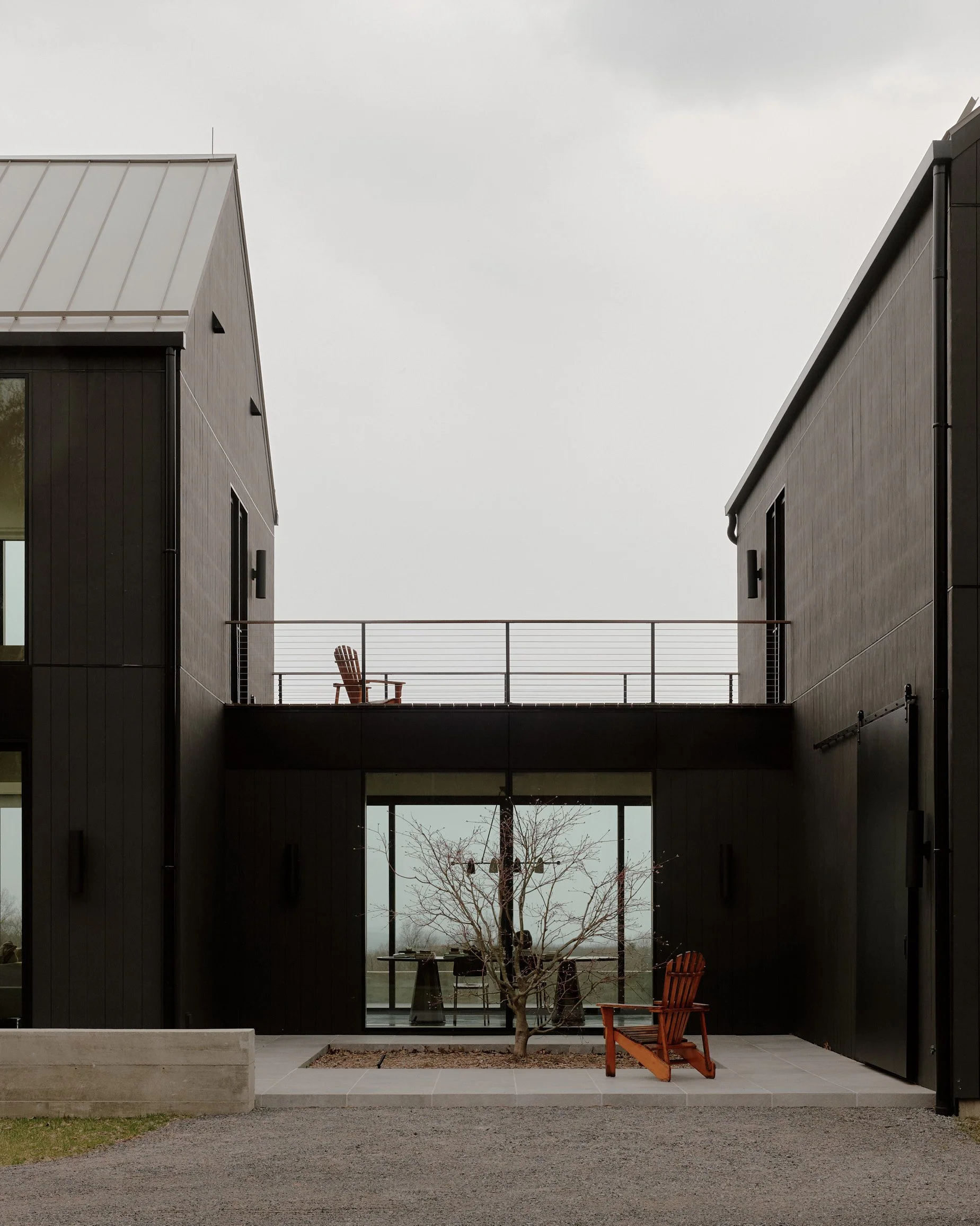 Courtyard between two black-clad gabled volumes joined by an upper walkway at a Ghent, NY home by JG Neukomm Architecture