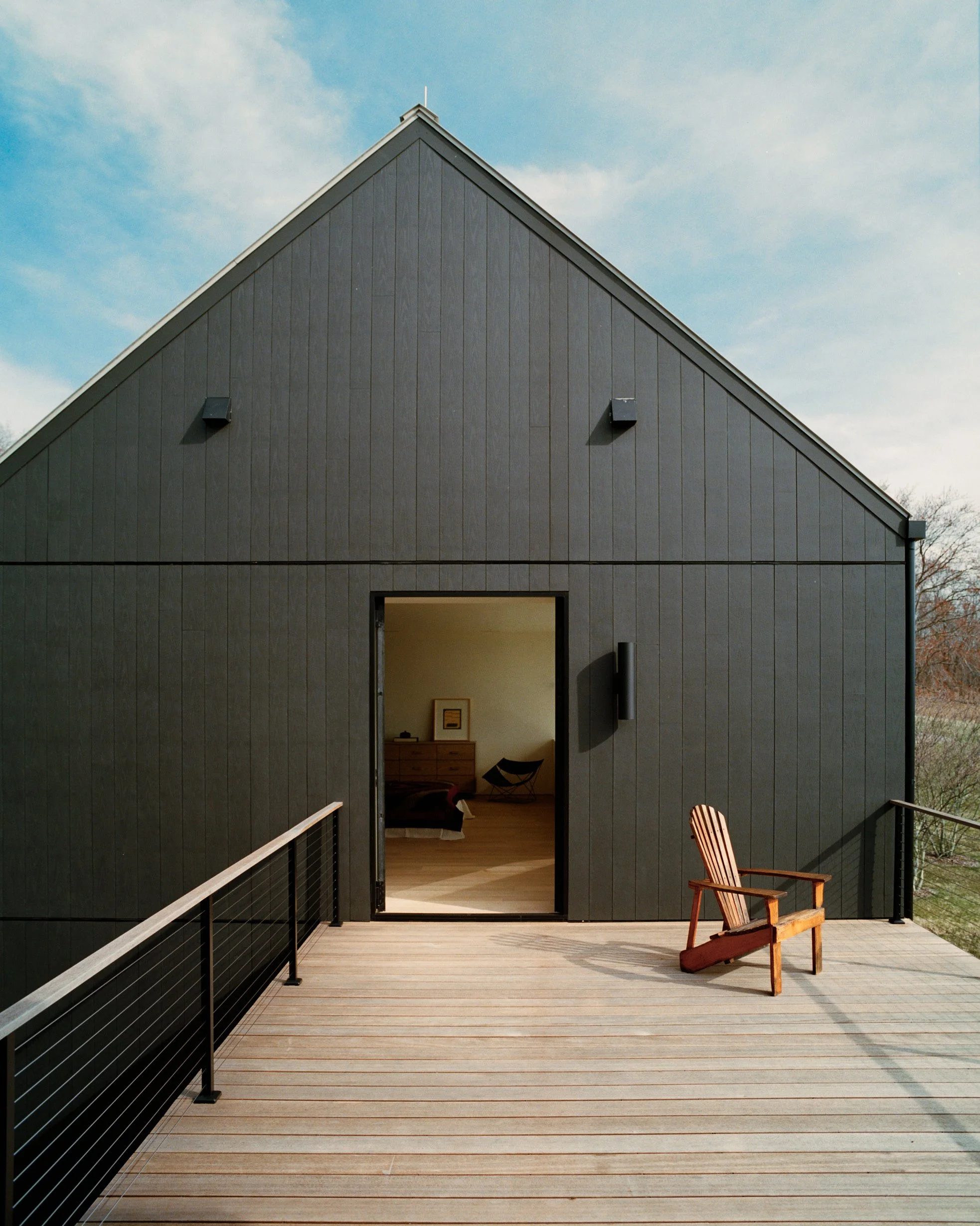 Black board-clad gabled exterior with a wood deck and cable railing at a Ghent, NY home by JG Neukomm Architecture