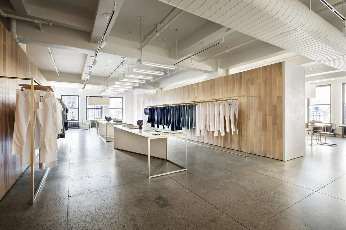 Wide view of the FRAME Denim showroom with an oak feature wall, central display table, and city windows, designed by JGNA