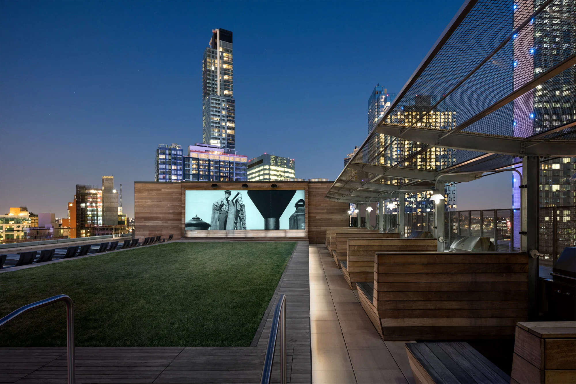 JGNA residential rooftop with a lawn, an outdoor projection screen, cabana seating, and a lit skyline at night