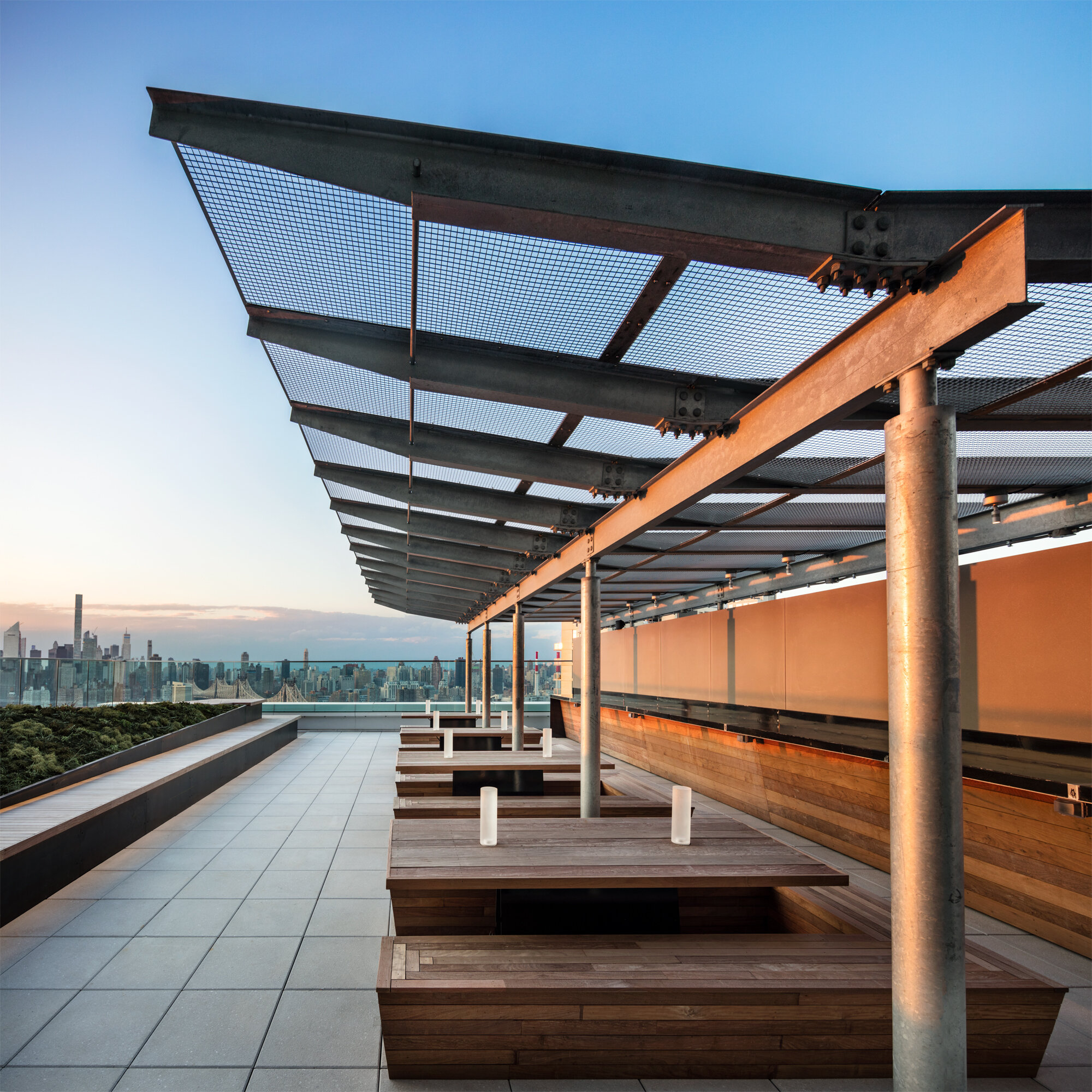 JGNA residential rooftop terrace with a steel mesh trellis, wood benches, and a city skyline at dusk