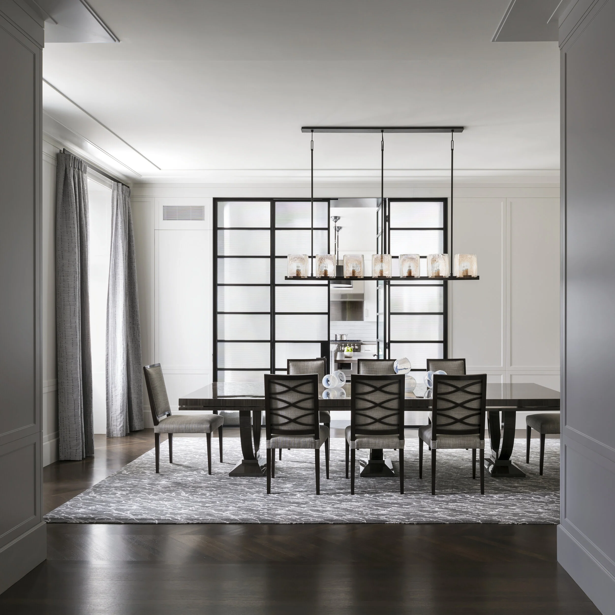 JGNA residence dining room with a long table, linear glass chandelier, and a steel-and-glass partition