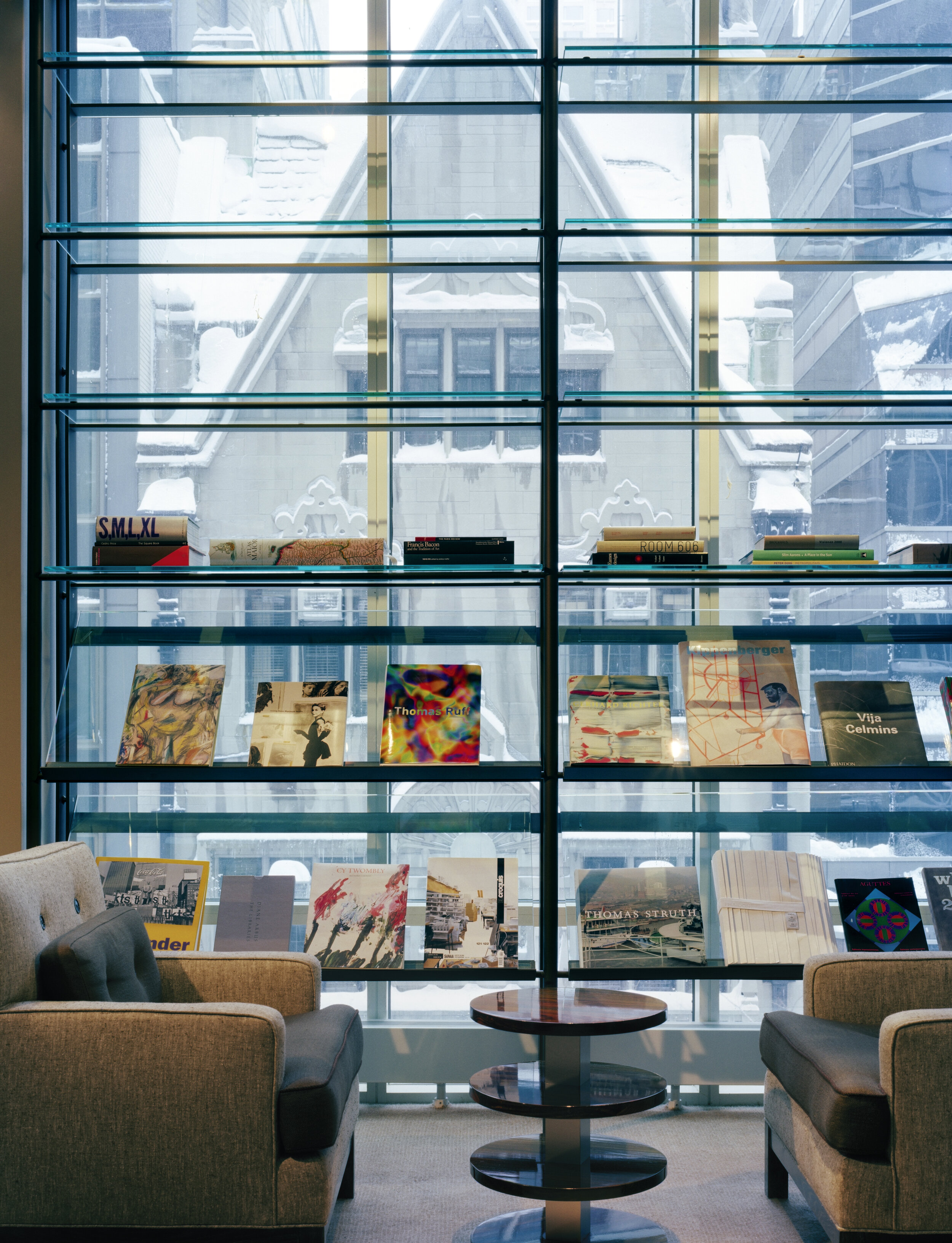 JGNA library with glass bookshelves against a window wall, paired armchairs, and a tiered side table